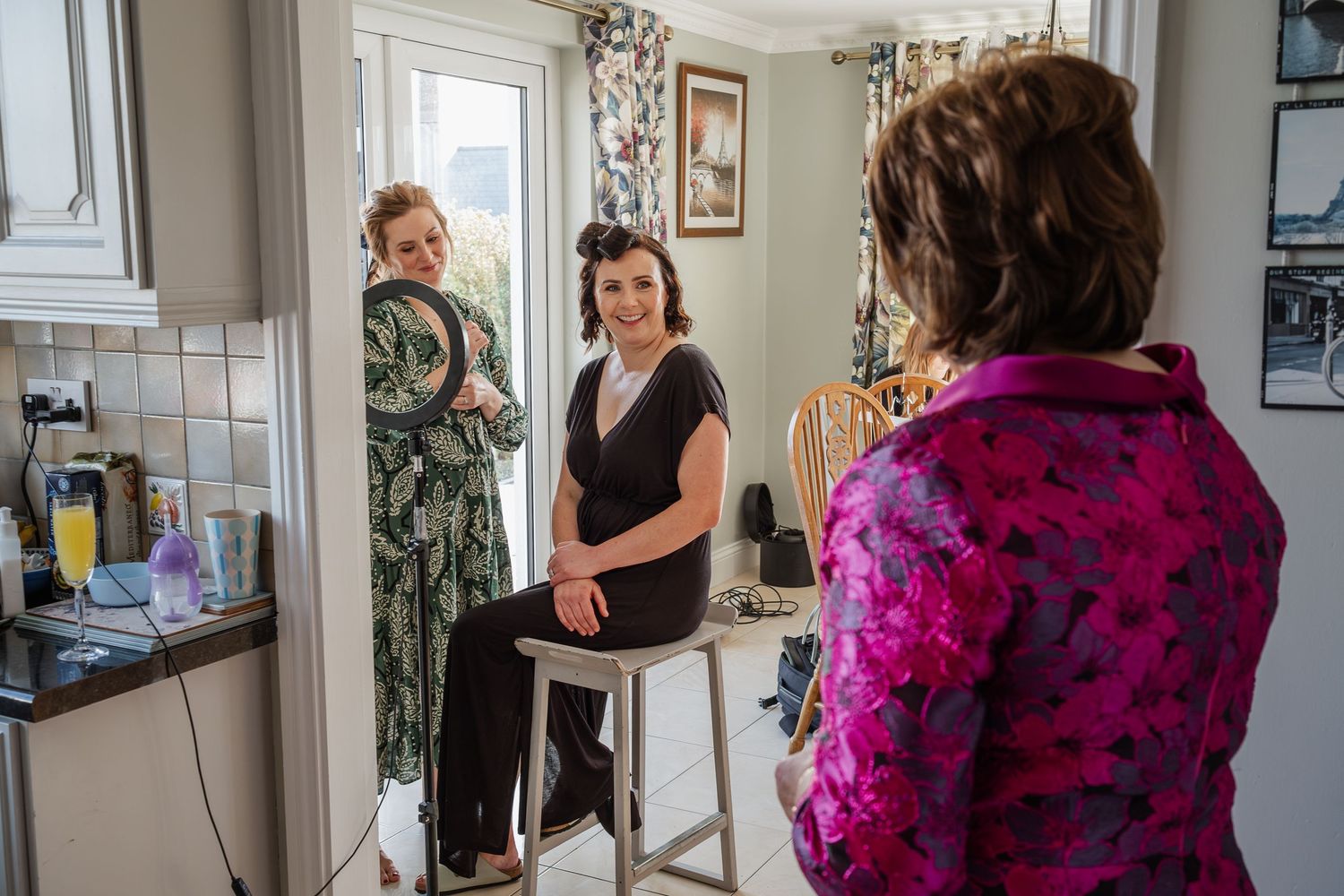 Three women chatting in a kitchen during bridal preparations, one seated on a stool wearing black outfit.