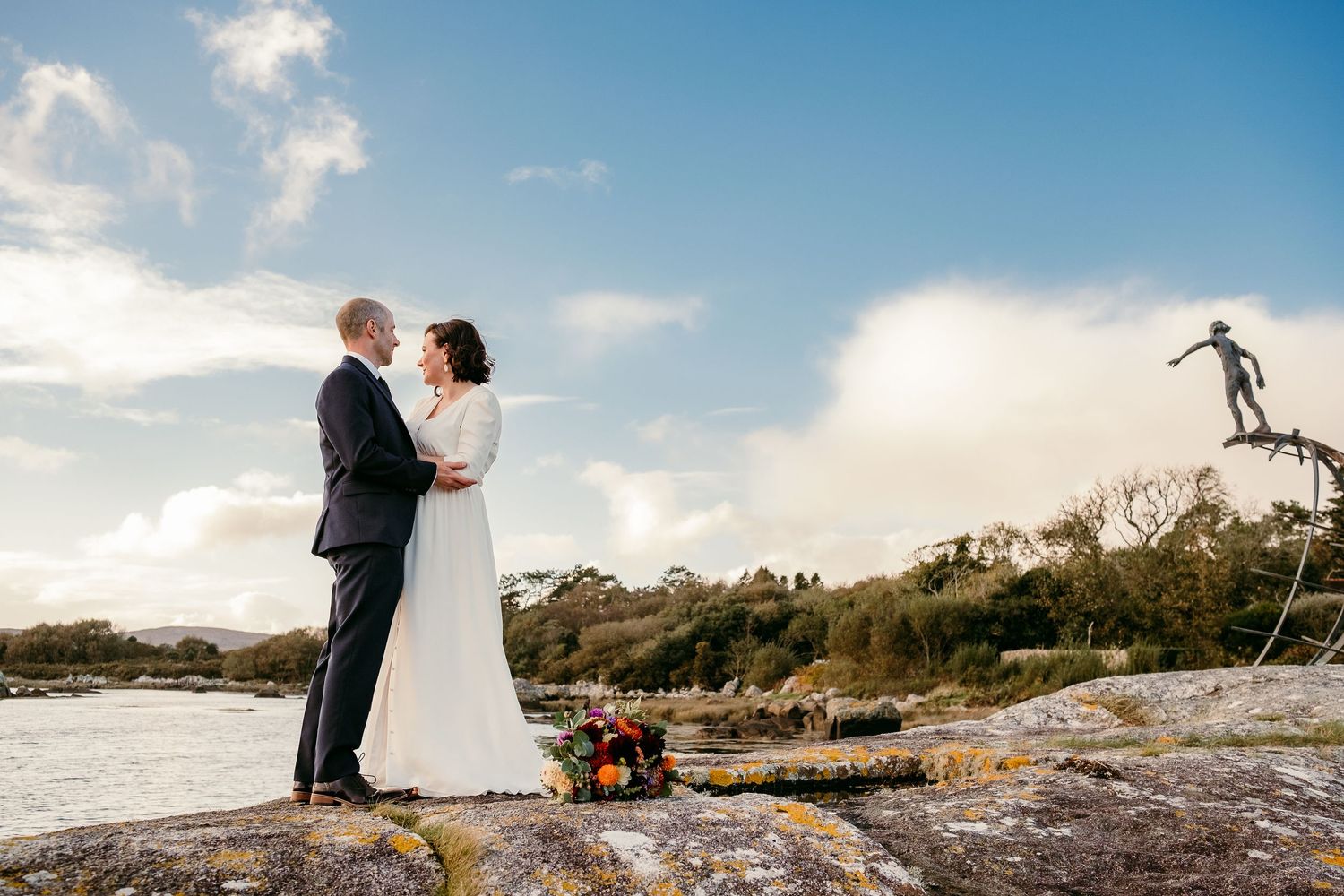 Bride and groom share a romantic moment on rocky coastal landscape with a bronze statue and blue skies in background.