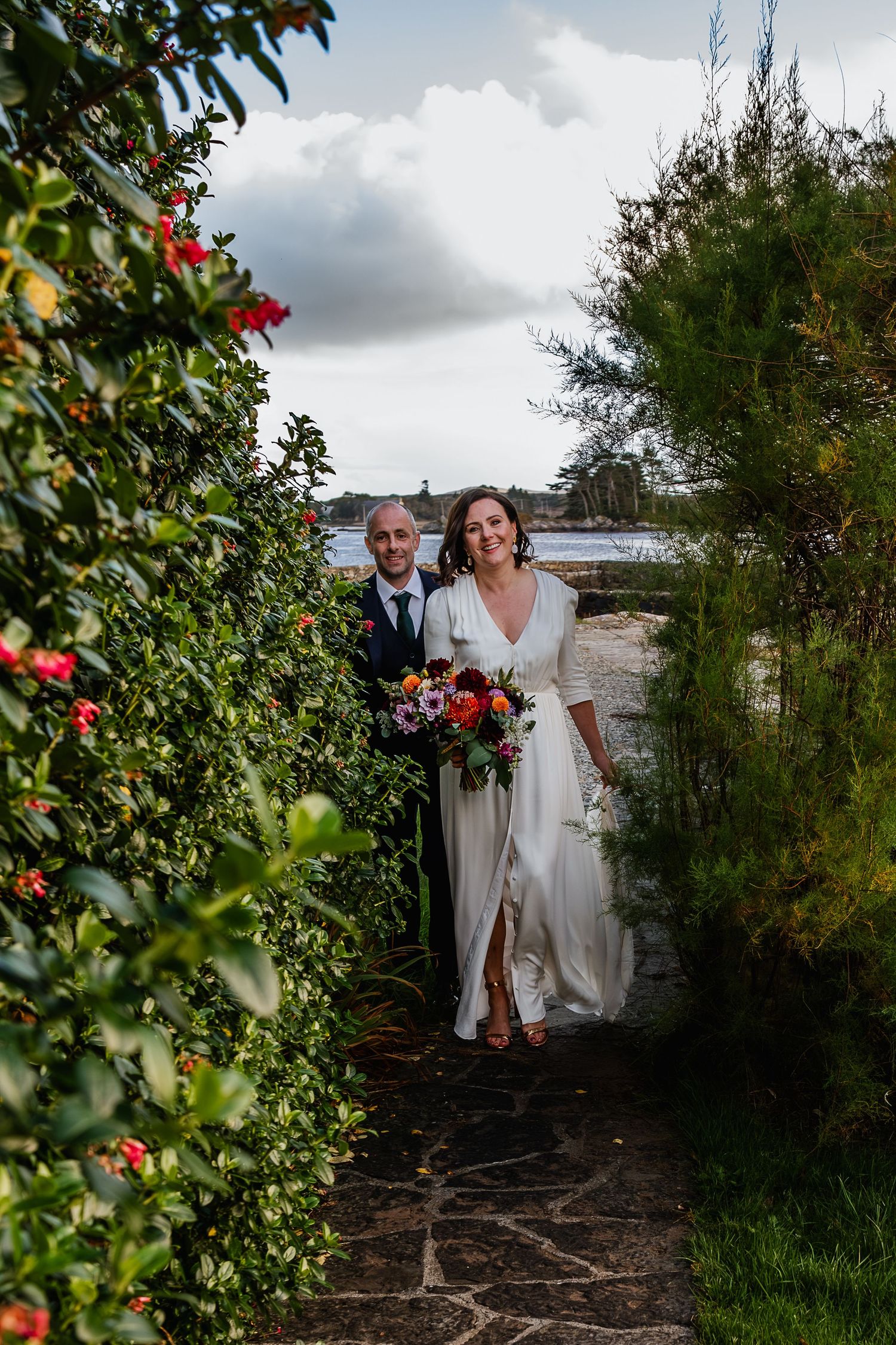 Bride and groom walking through lush garden pathway with colorful bouquet, water visible in background.