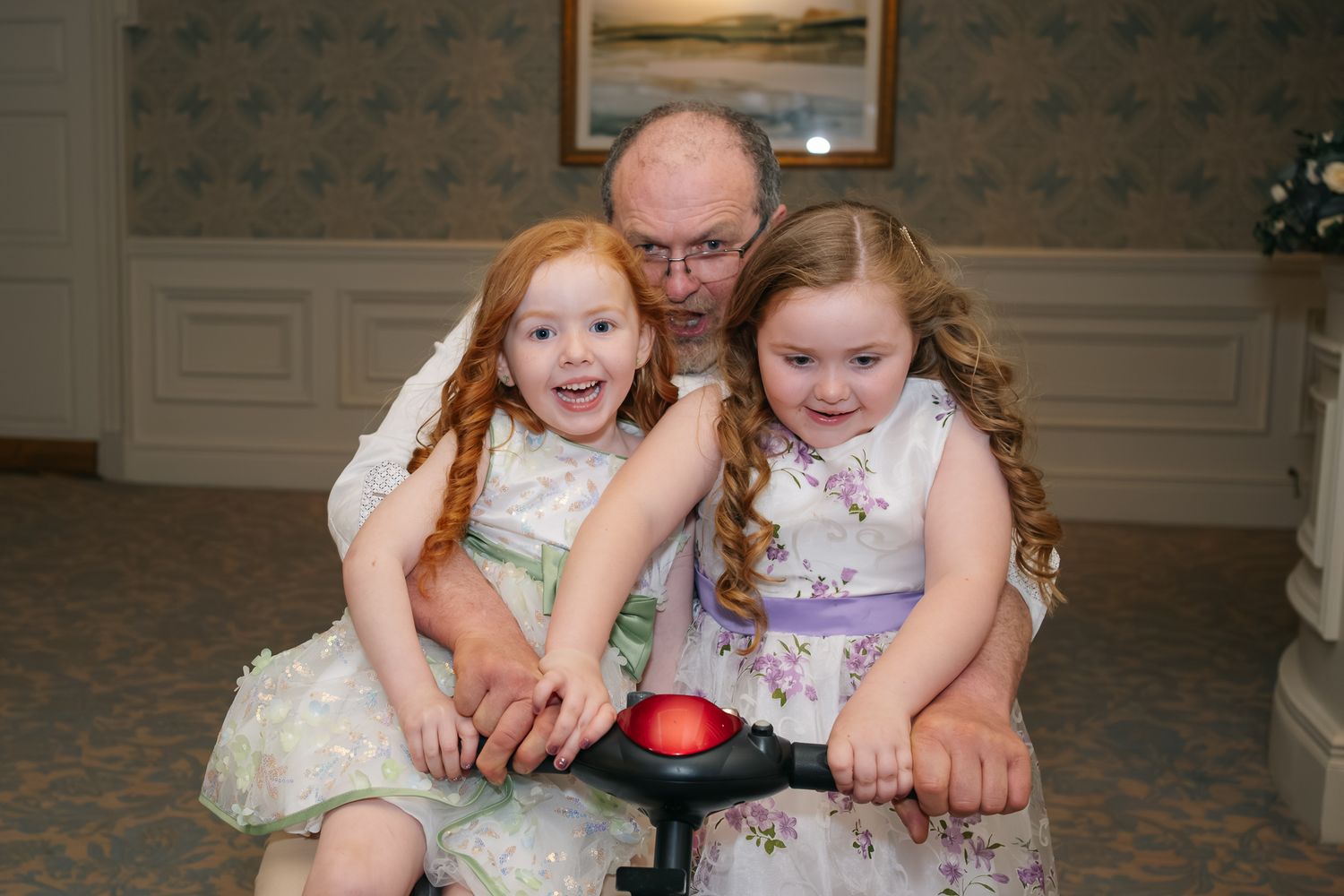 Grandfather smiling with two young girls in floral dresses sitting on a toy bike at a wedding reception.