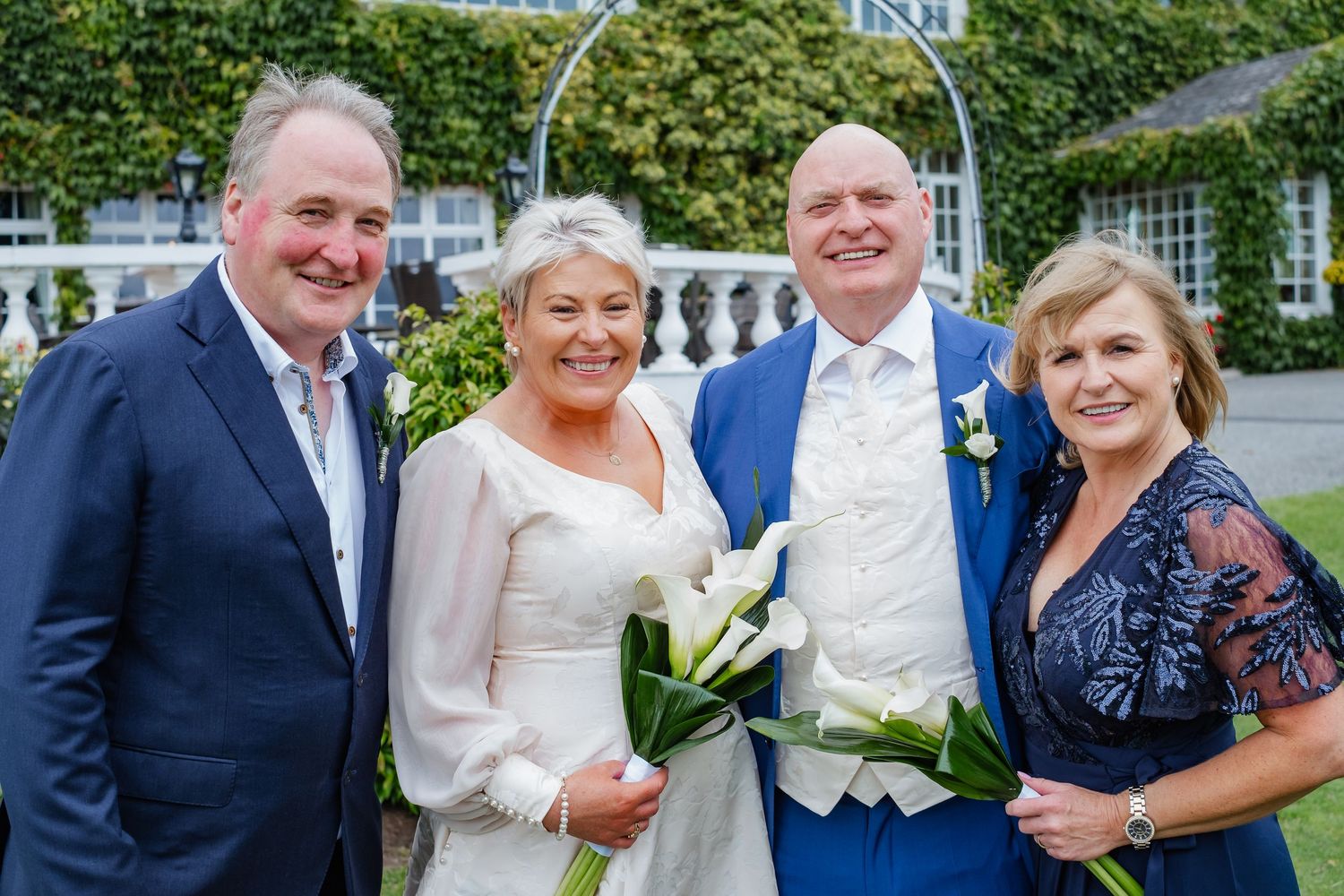 Wedding group photo of bride and groom with two guests outside ivy-covered venue.