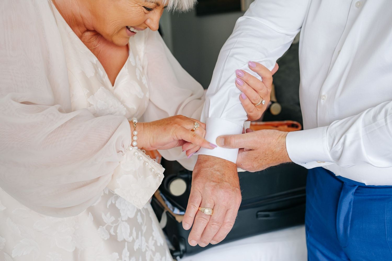 Woman helping groom fasten cufflinks on white shirt before wedding ceremony.