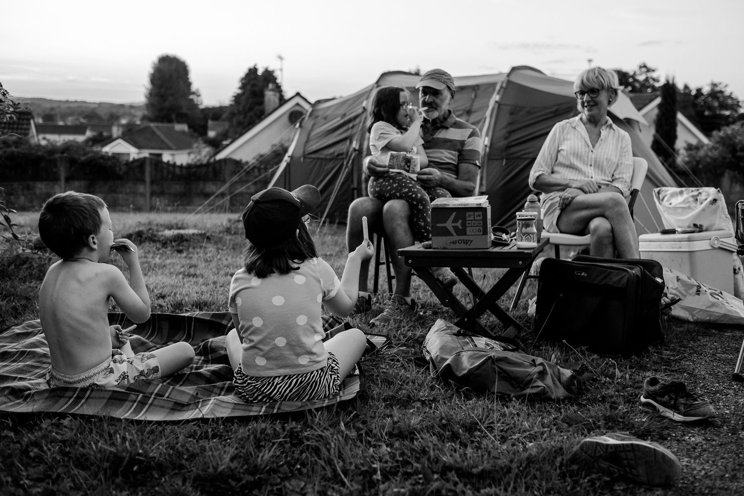 Black and white photo of a family camping outdoors, sitting on grass near a tent, sharing a meal together.