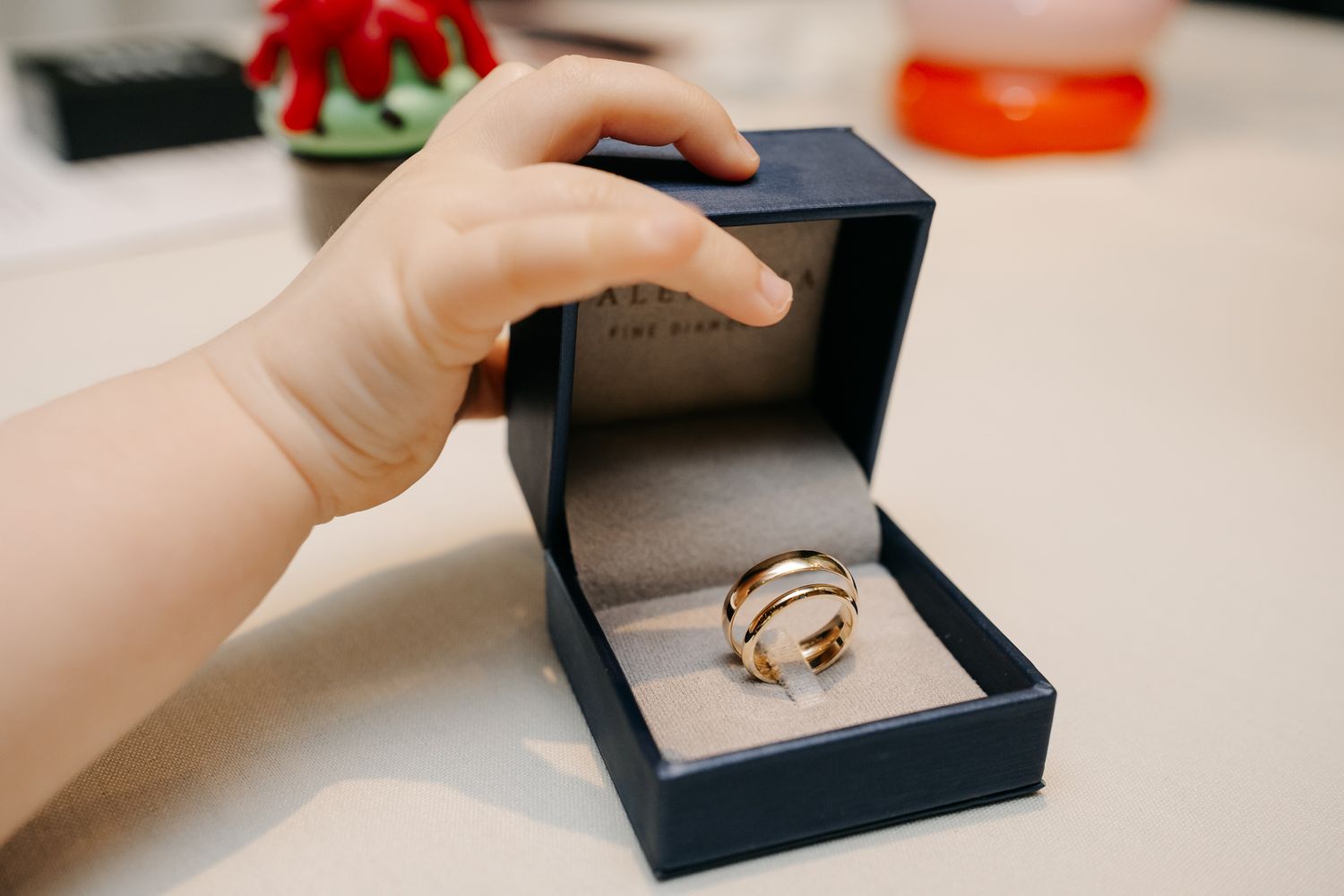Child's hand reaching for a gold trinity ring in a navy blue jewelry box on a white surface.