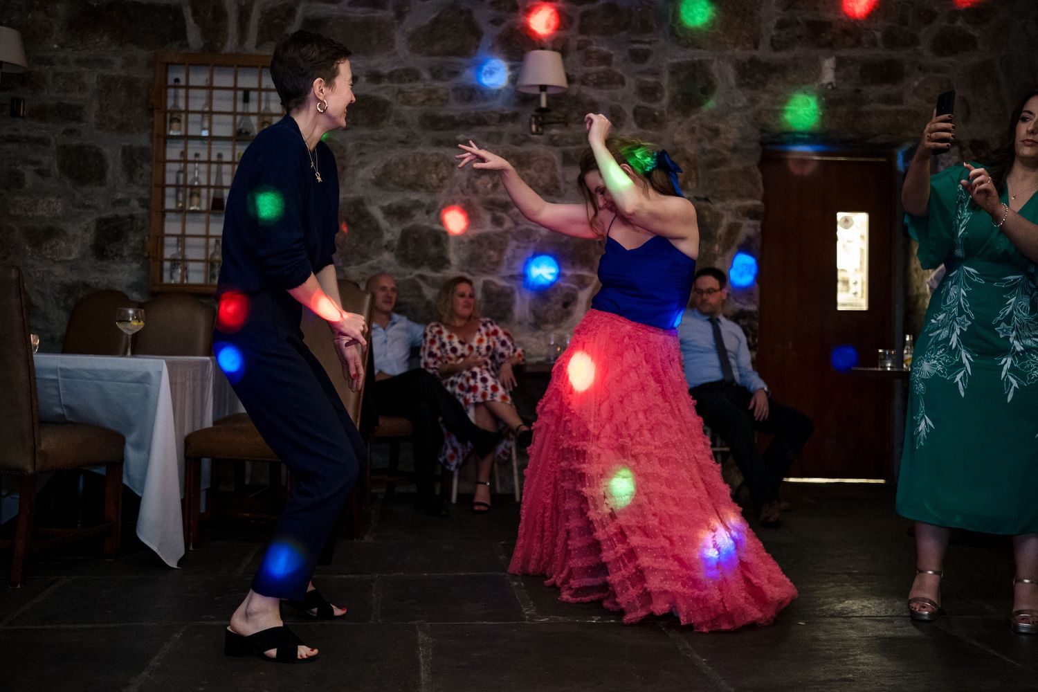 Couple dancing energetically with colorful bokeh lights at a festive wedding reception in a stone venue.