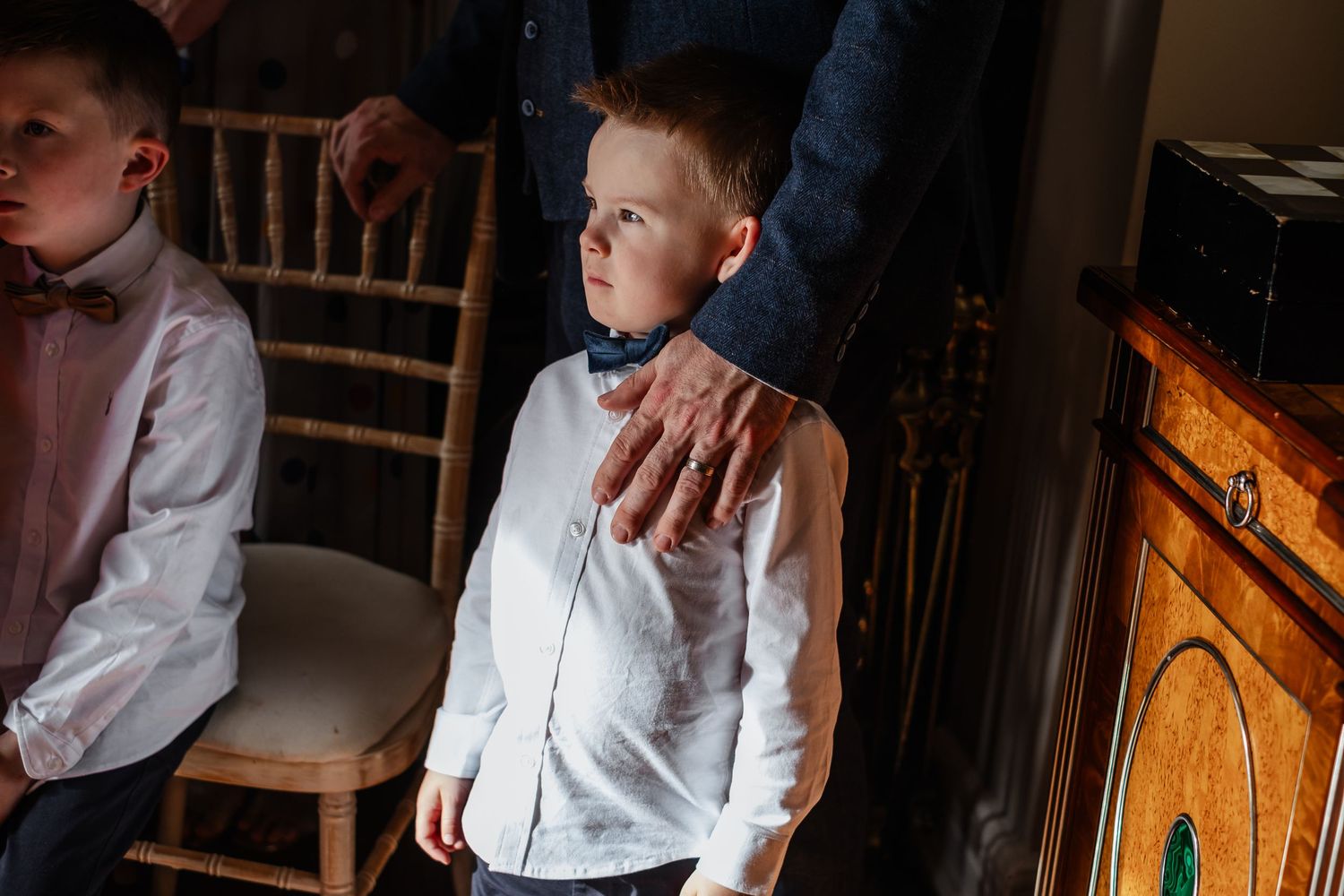 A young boy in a white shirt and blue bow tie stands attentively as an adult adjusts his collar at a wedding.