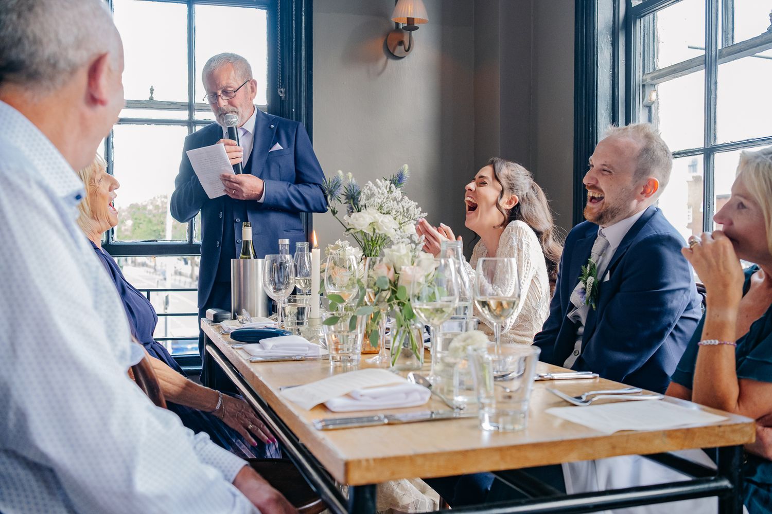 Wedding guests enjoying a meal and conversation at a reception dinner in a bright restaurant setting.