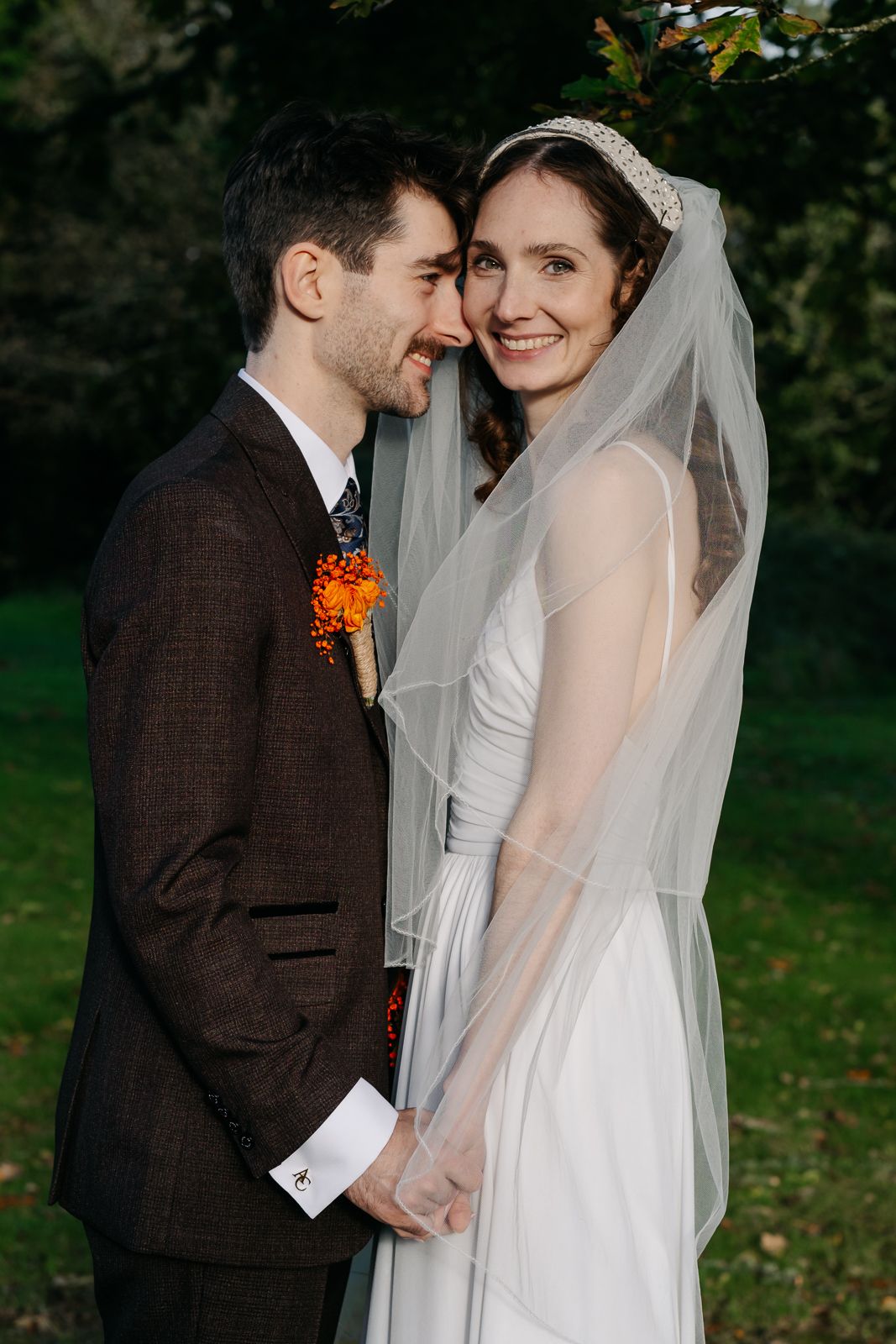 Smiling bride in white gown and veil holds hands with groom in brown suit with orange boutonniere outdoors.