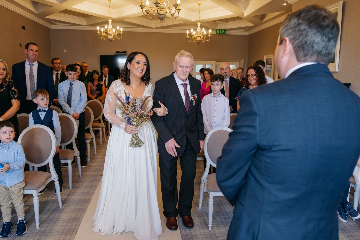 Bride walks down the aisle with escort during indoor wedding ceremony, guests watching in elegant venue.