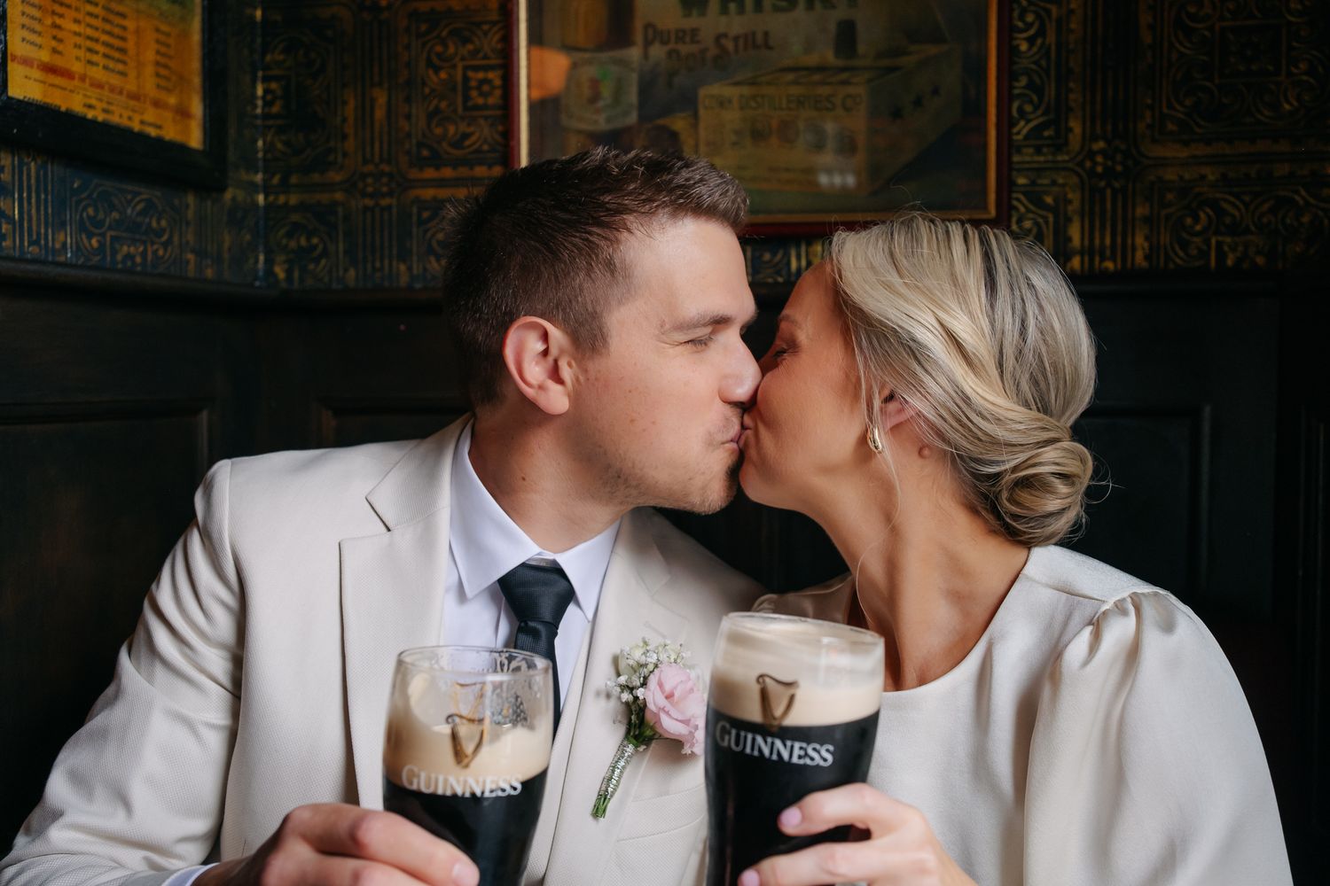 Newlywed couple kissing while holding Guinness pints in a cozy pub, bride in white dress and groom in ivory suit.