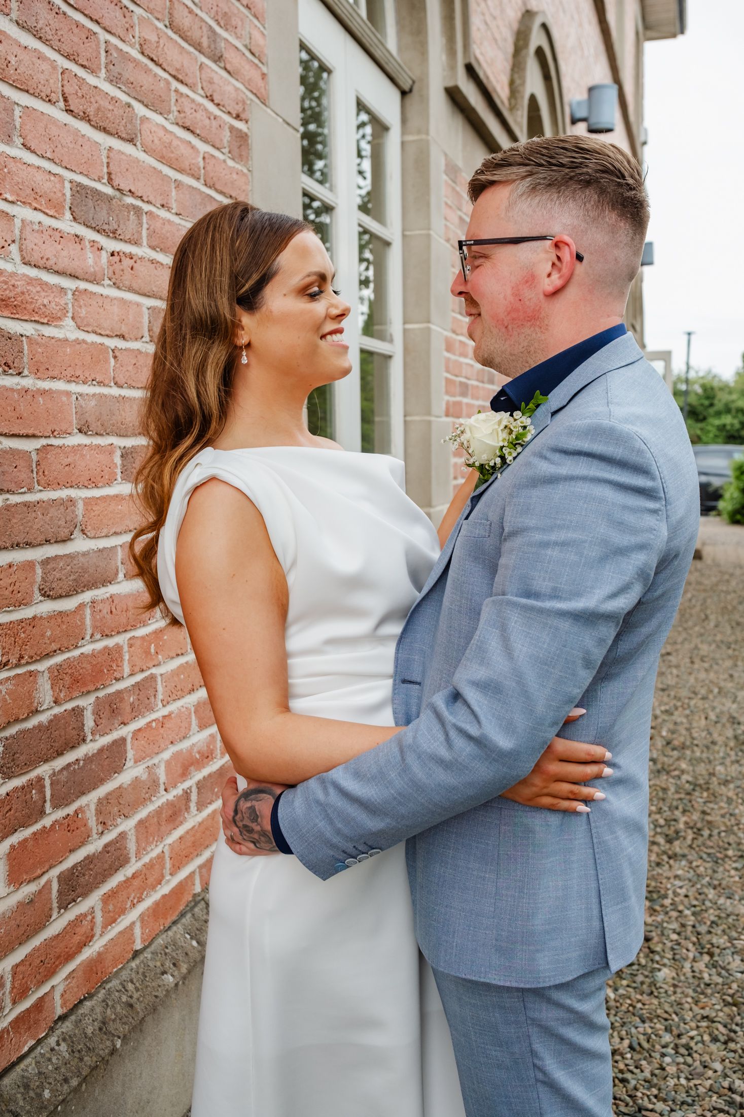 Wedding couple poses against rustic brick wall in elegant attire.