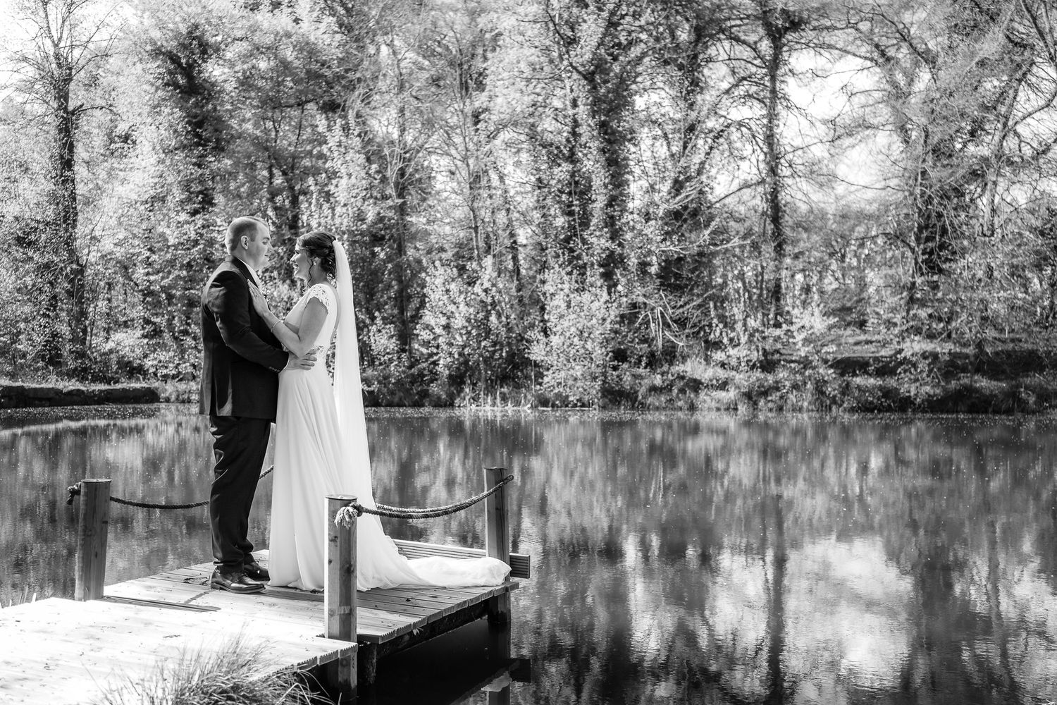 Black and white photo of a wedding ceremony by a serene pond with trees in the background.