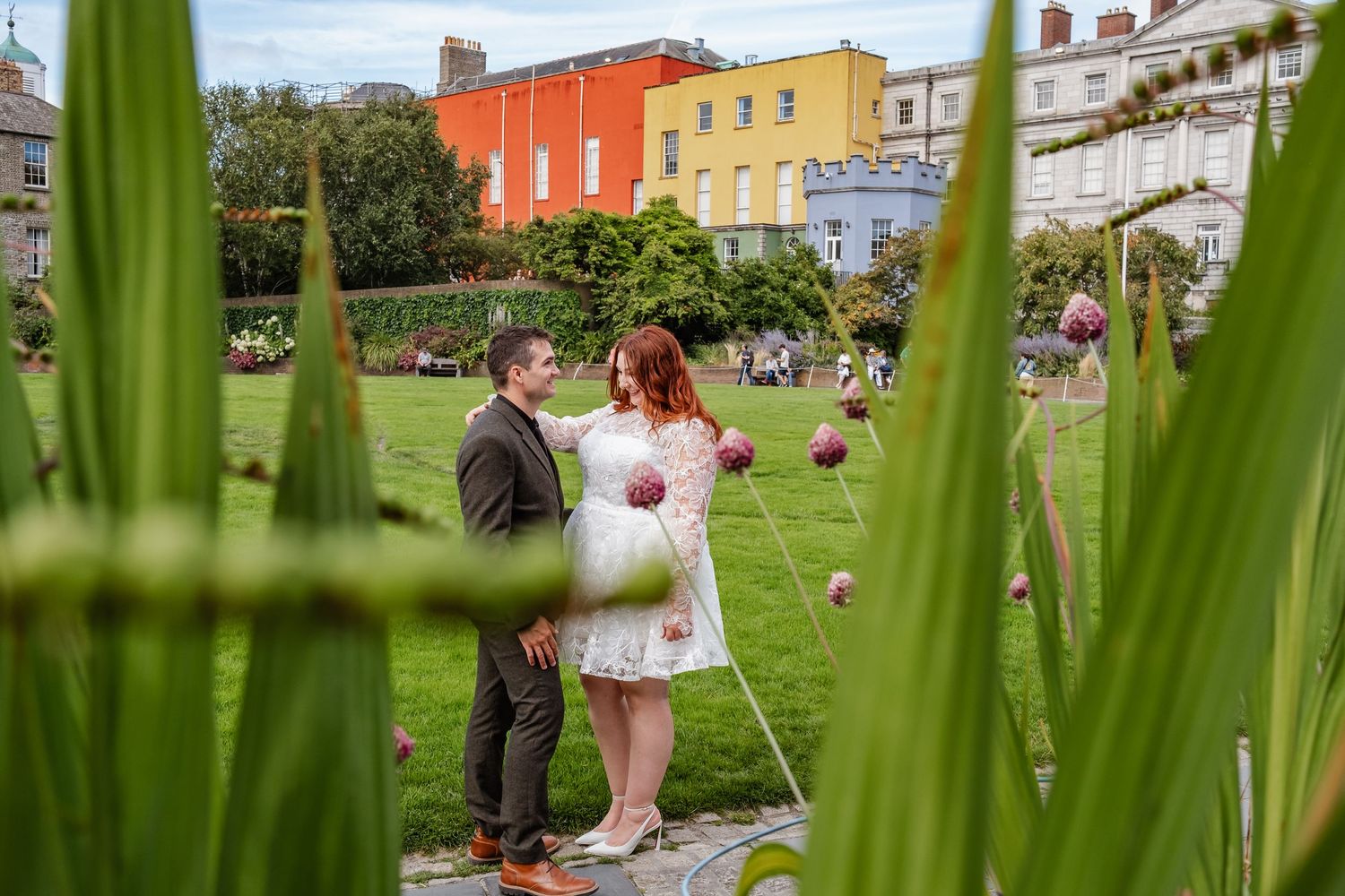 A couple photographed through green foliage against colorful buildings in an urban garden setting.