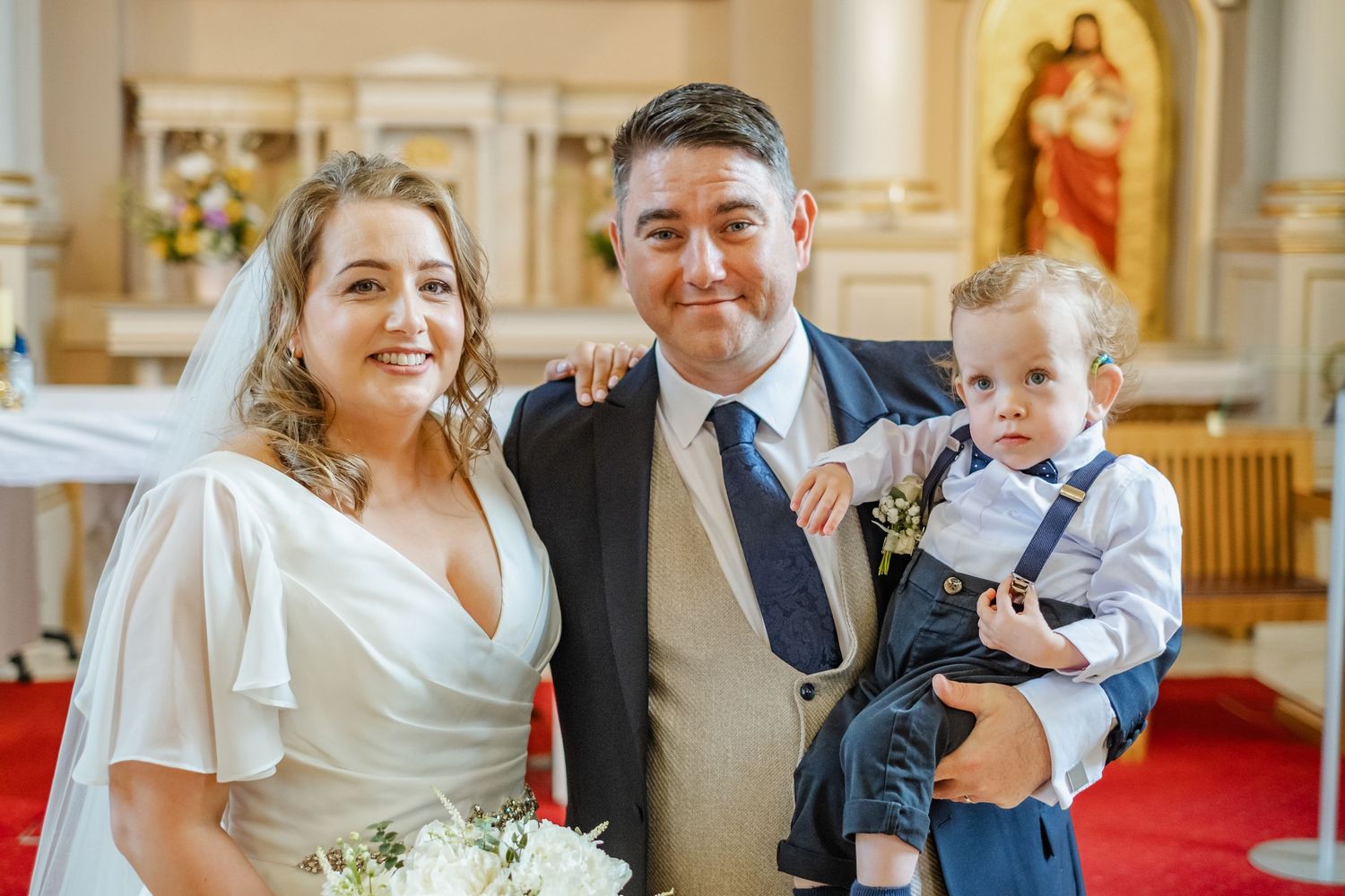 Newlyweds and small child pose for photo inside ornate church with religious artwork in background.