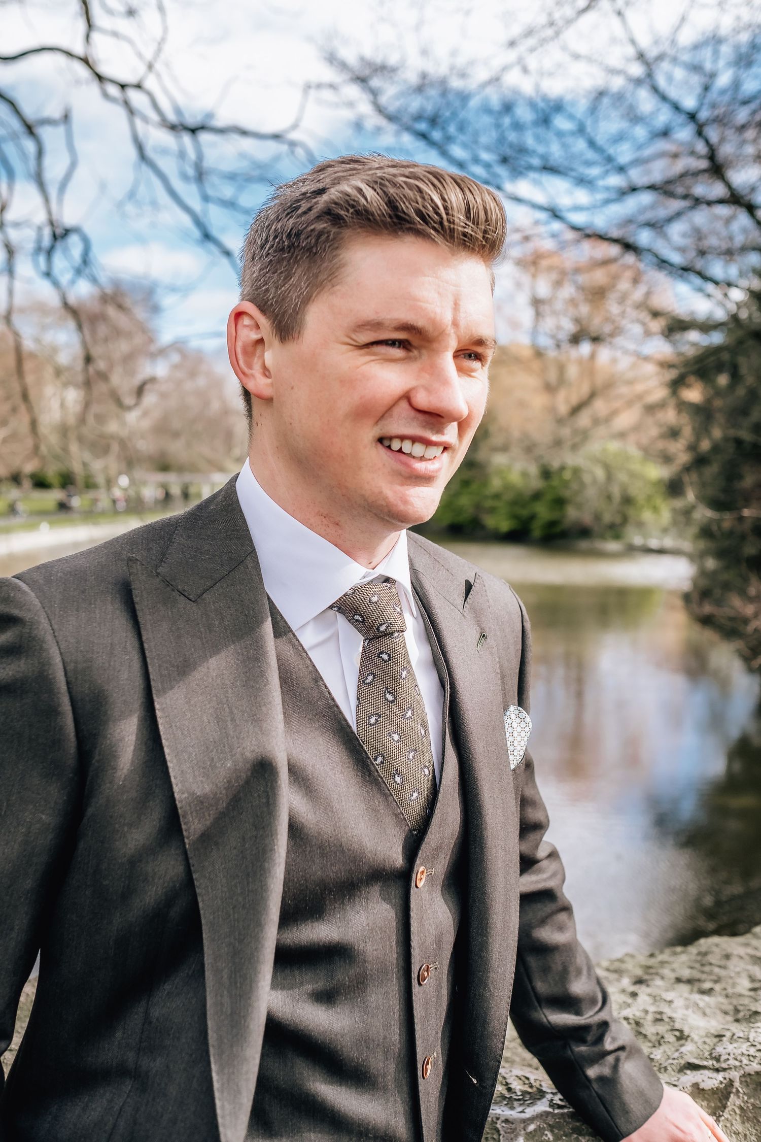 Groom in three-piece suit poses by pond in park setting.