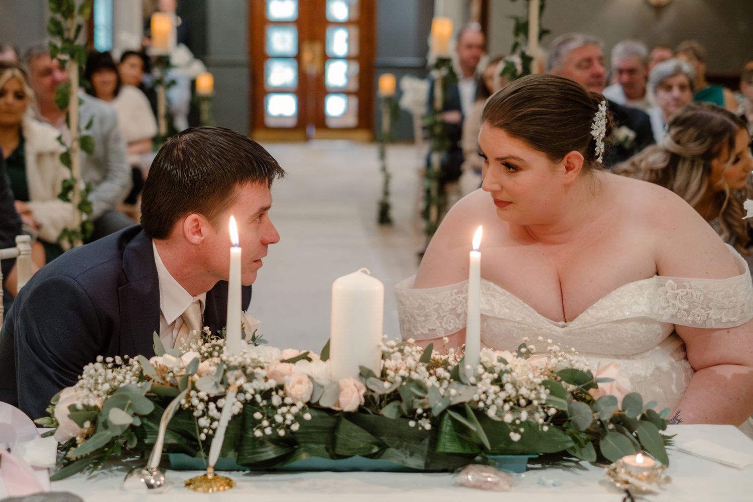 Couple seated at decorated wedding table with lit unity candles and greenery centerpiece.