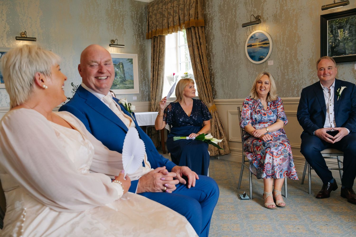 Bride and groom laughing with wedding guests during intimate ceremony in elegant room.