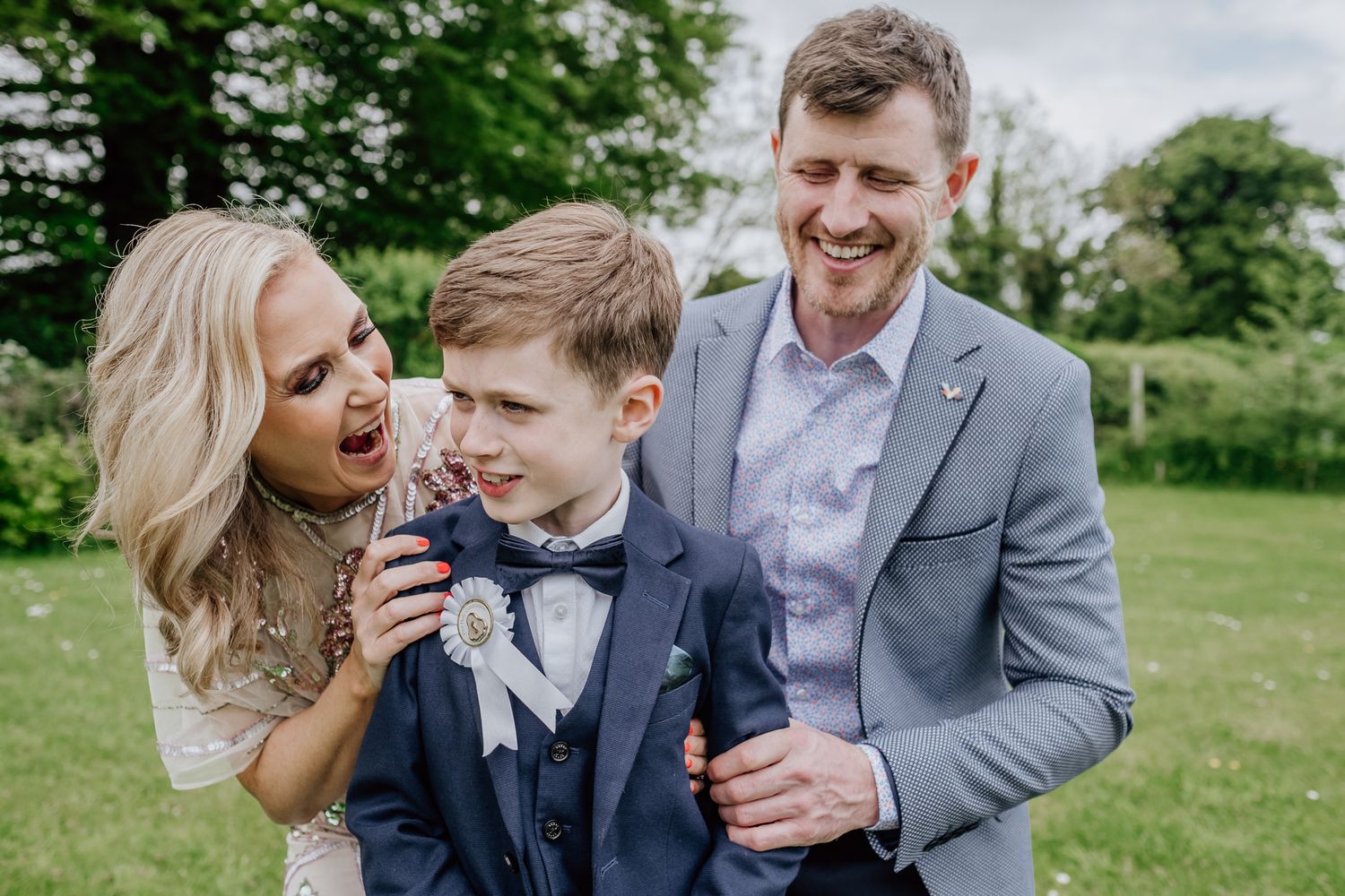 A joyful family portrait with a blonde woman, young boy in navy suit, and man in gray suit laughing outdoors.