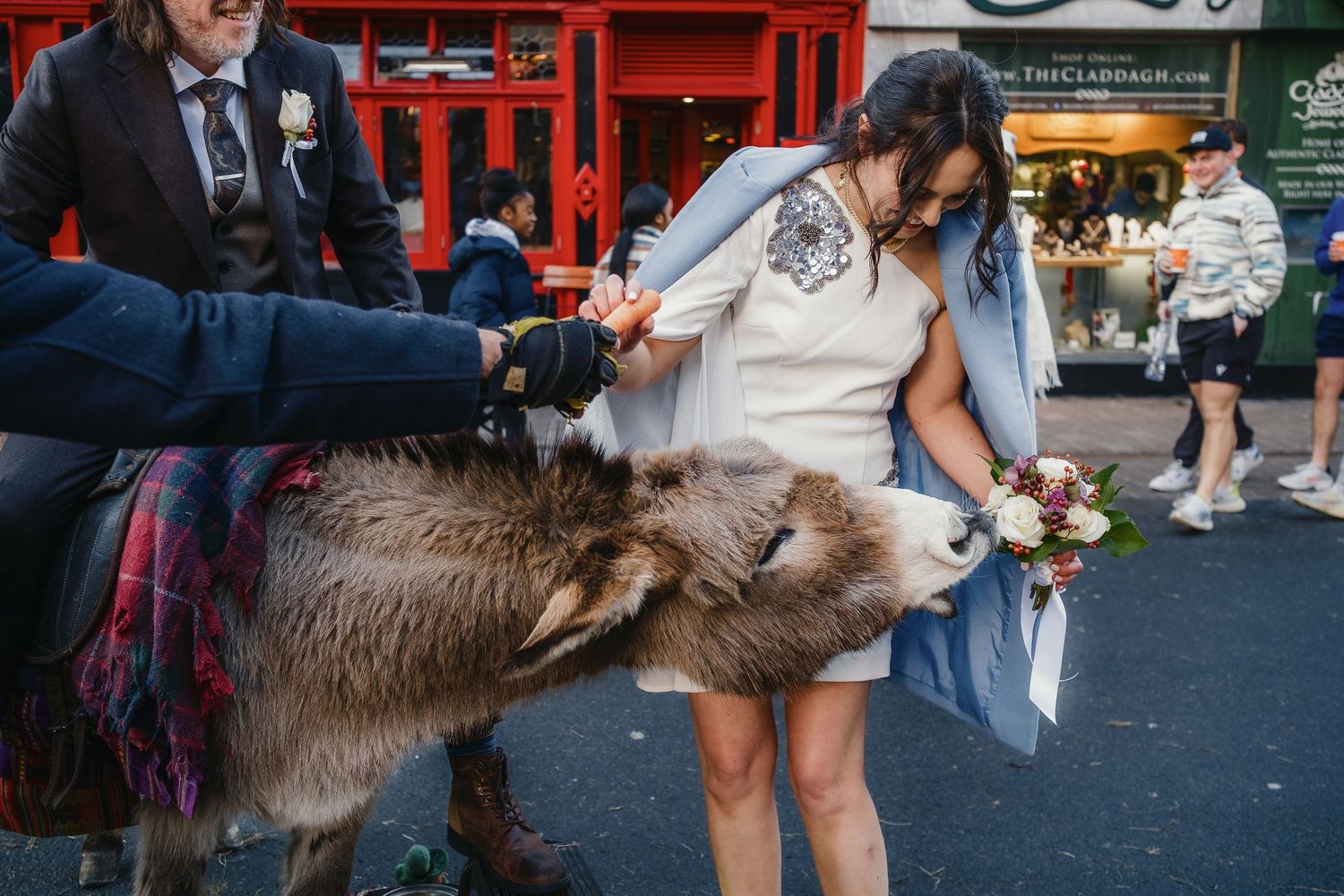 A bride in a white dress holds flowers while a donkey nuzzles her on a busy street during a wedding.