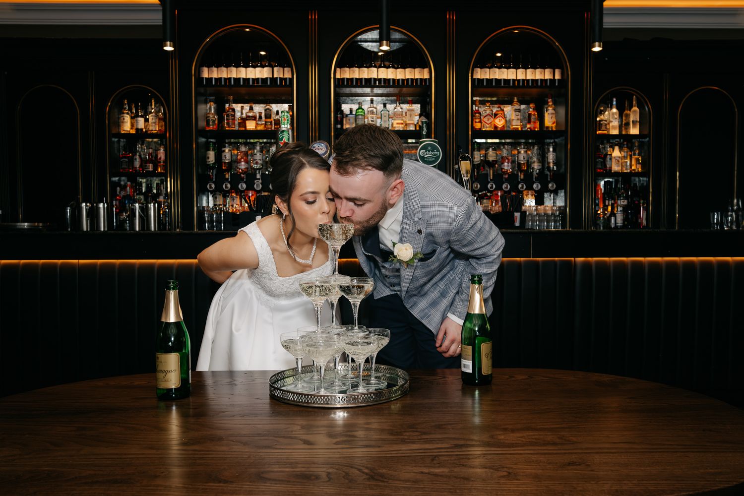Bride and groom kissing over a champagne tower at a bar, with bottles of champagne on the counter.