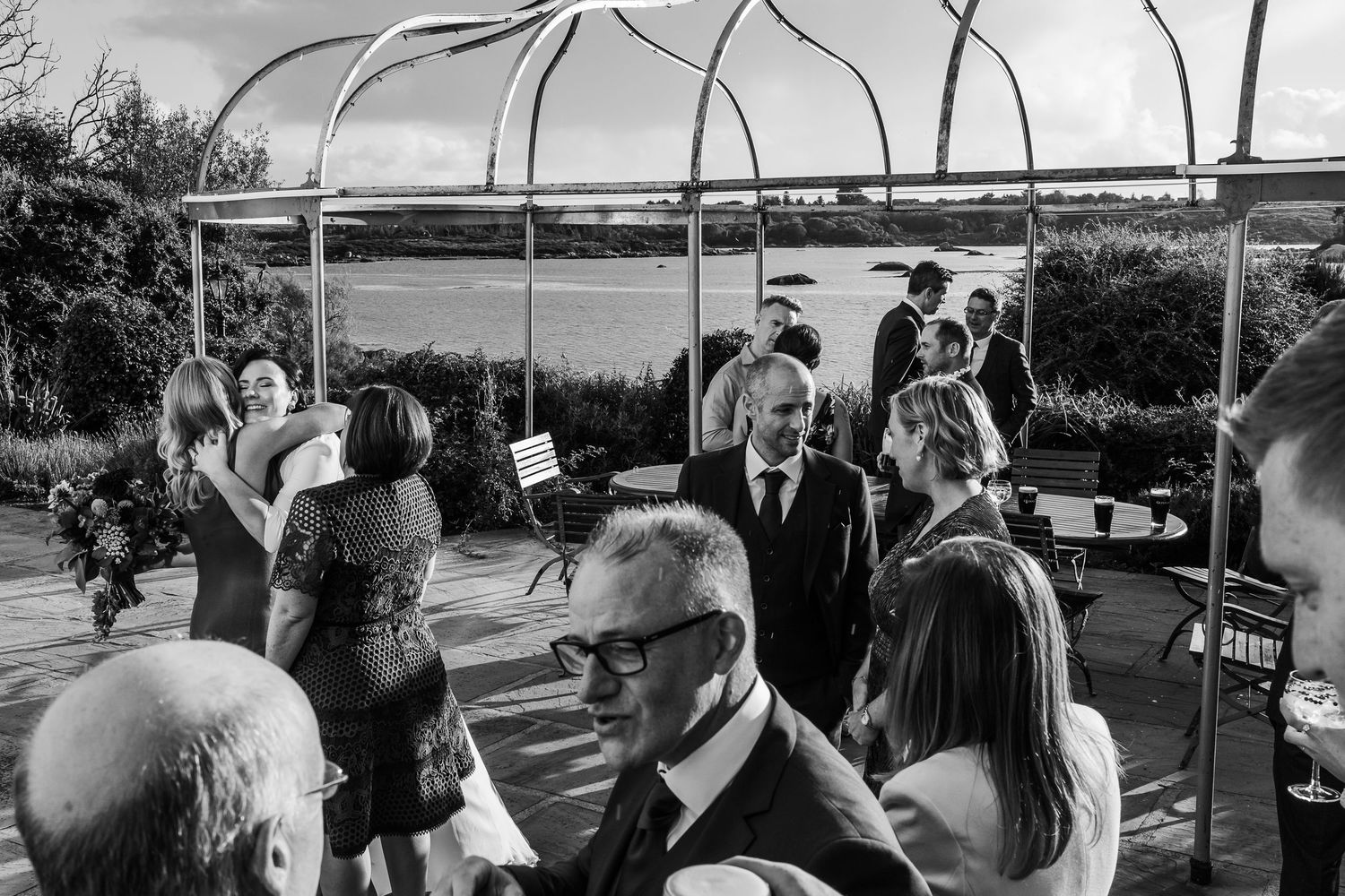 Black and white photo of wedding guests mingling on an outdoor terrace with a metal pergola and river views.