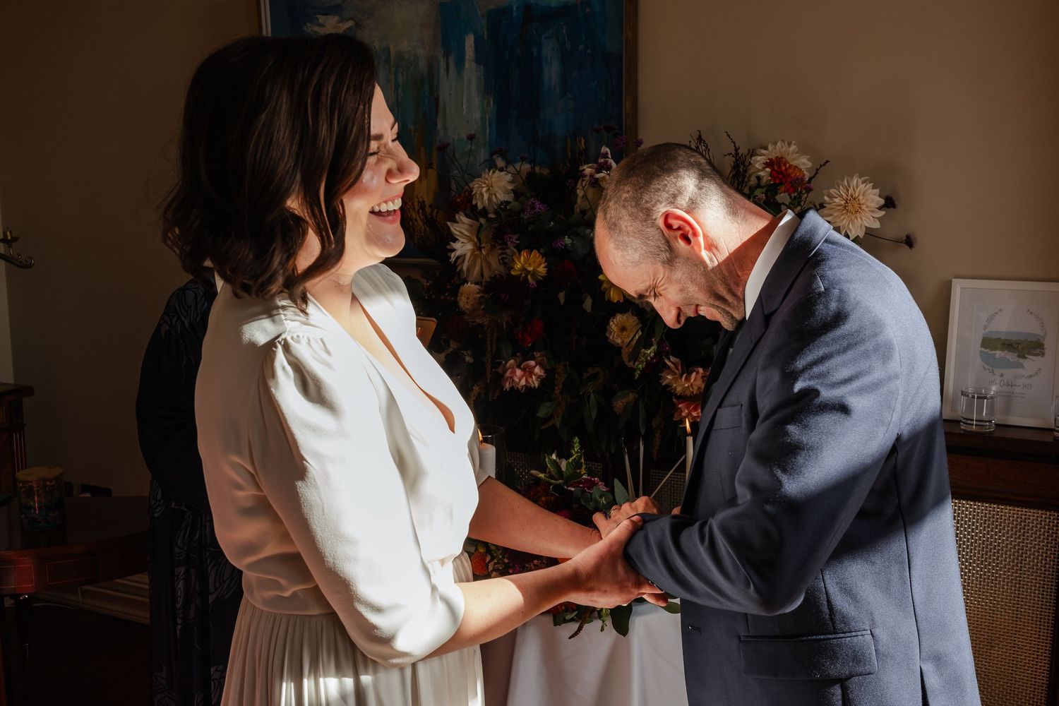 Bride and groom exchanging rings indoors, smiling joyfully, with colorful floral arrangement in background.