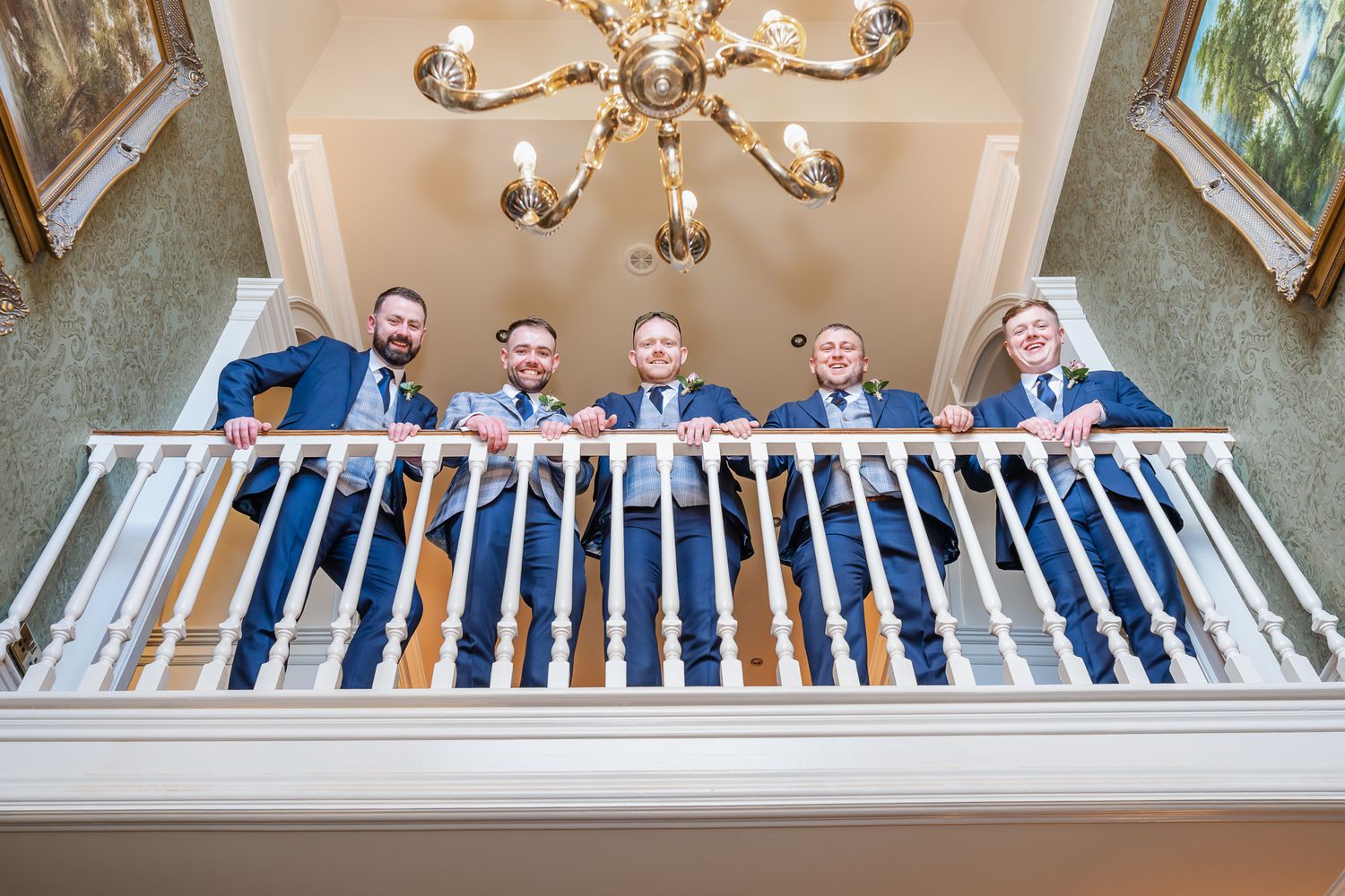 Five groomsmen in blue suits lean over a white balcony railing beneath a gold chandelier in an elegant venue.