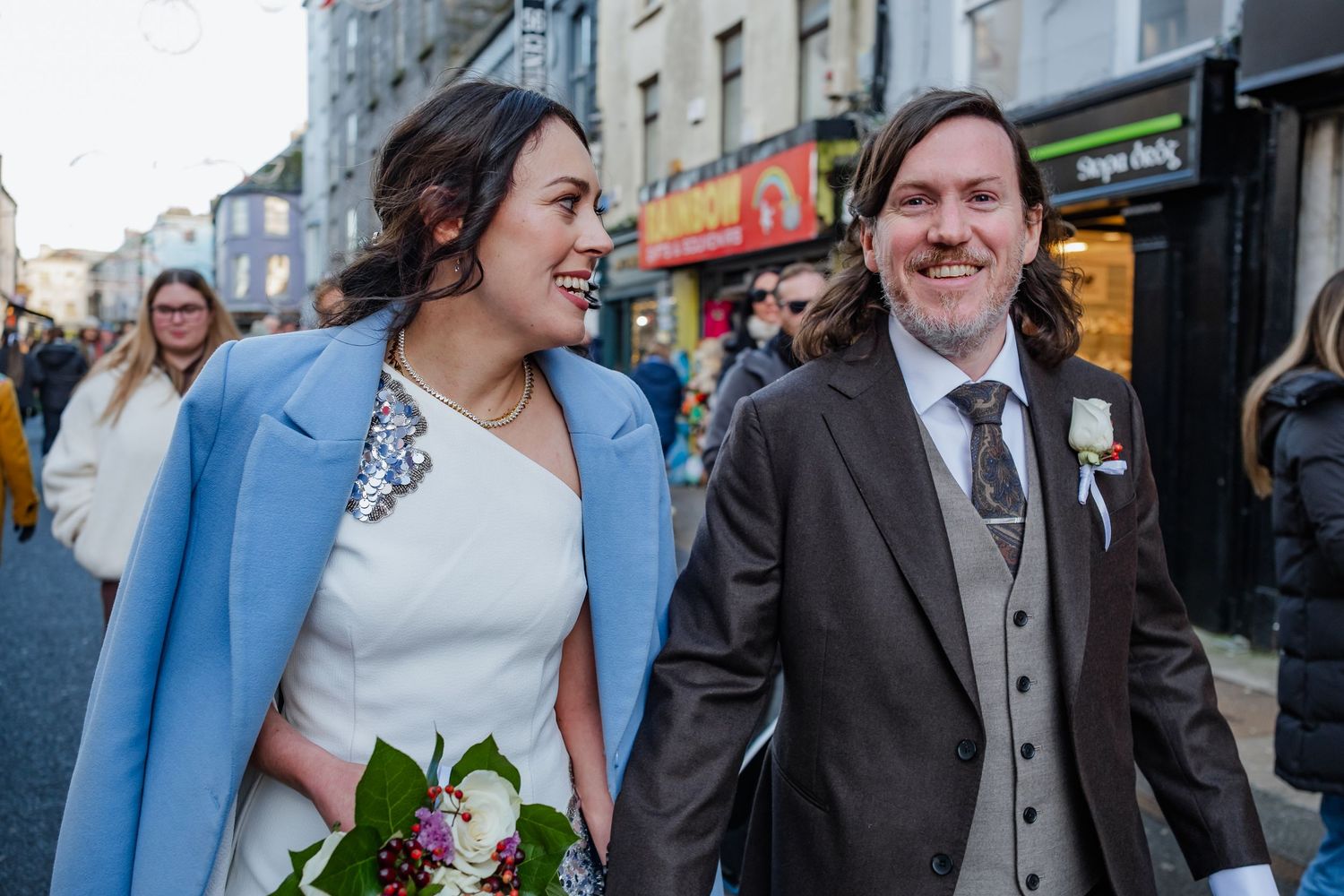 Happy newlywed couple walking down a busy street, bride in blue coat holding bouquet, groom in grey suit with boutonniere.