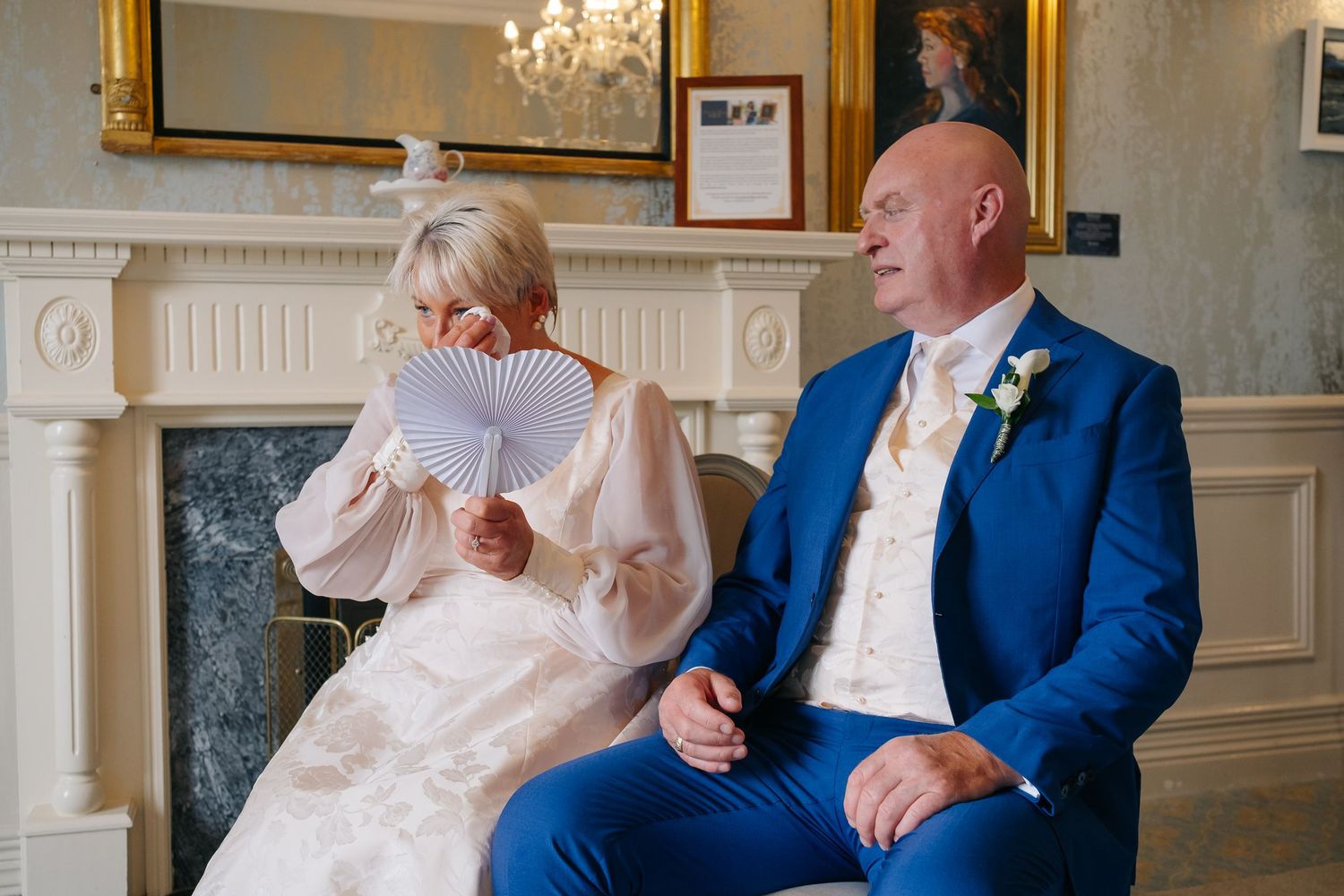 Bride fanning herself with white fan beside groom in blue suit during wedding ceremony.