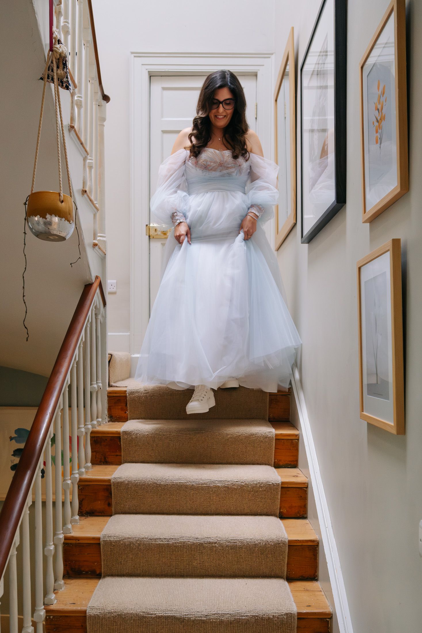 Bride in light blue tulle wedding dress and glasses descending wooden staircase in elegant home.