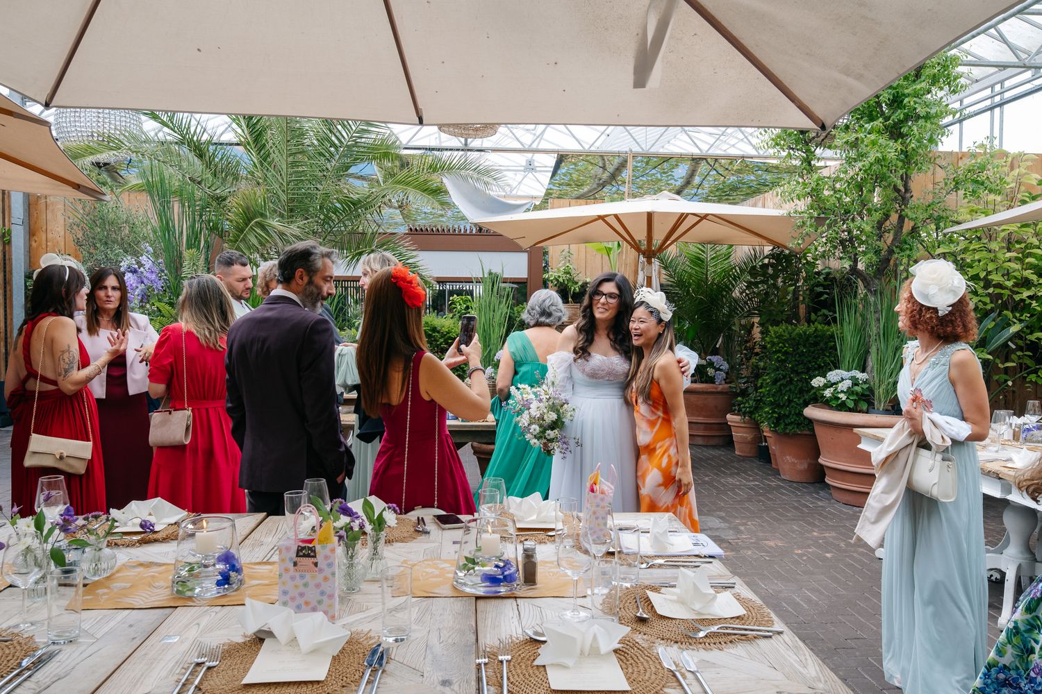 Elegantly dressed wedding guests mingle at an outdoor reception venue with lush greenery and decorated dining tables.