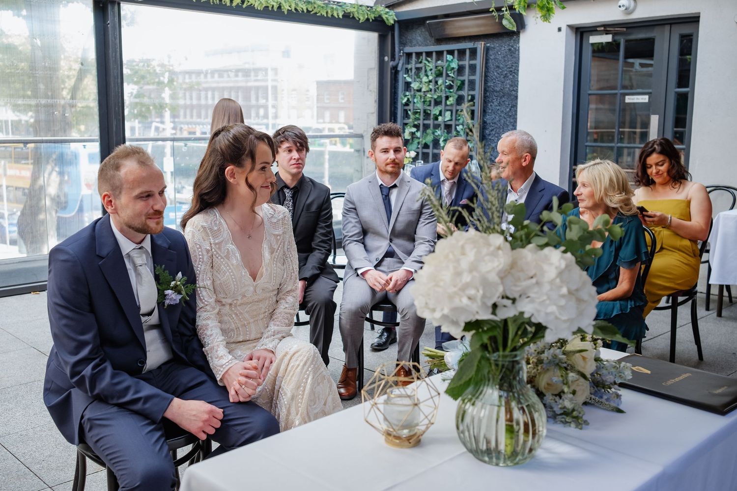 Wedding guests mingle and socialize at an elegant indoor reception with floral decorations and natural lighting.