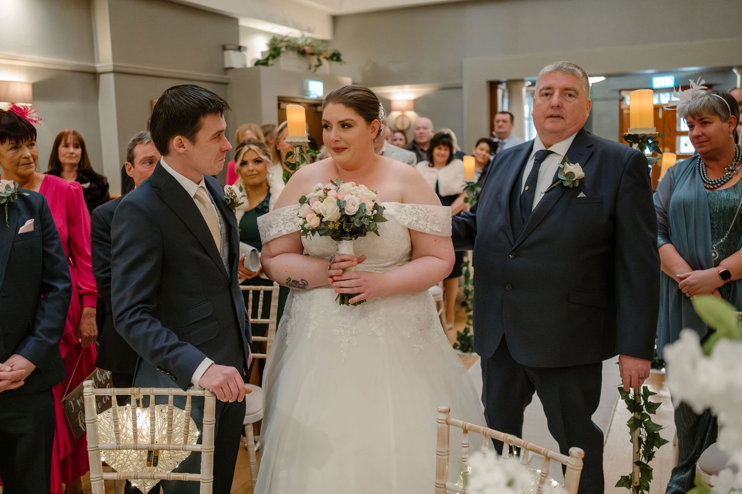 A wedding ceremony in progress in an elegant indoor venue with white chairs and guests looking on.