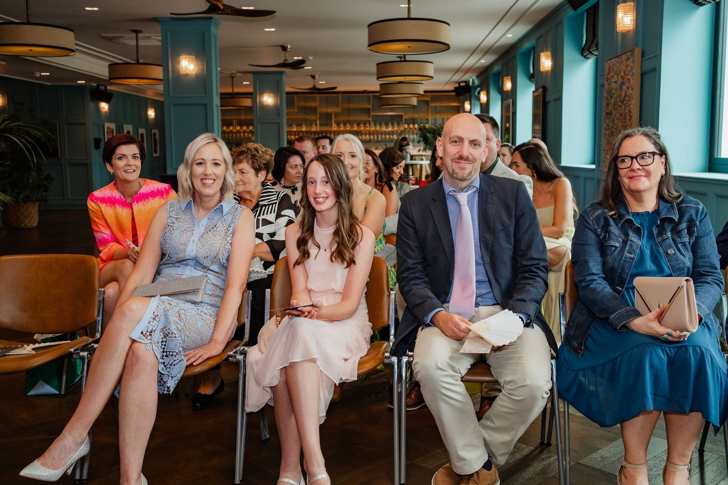 Wedding guests seated in rows at an intimate indoor ceremony with blue walls and warm lighting.