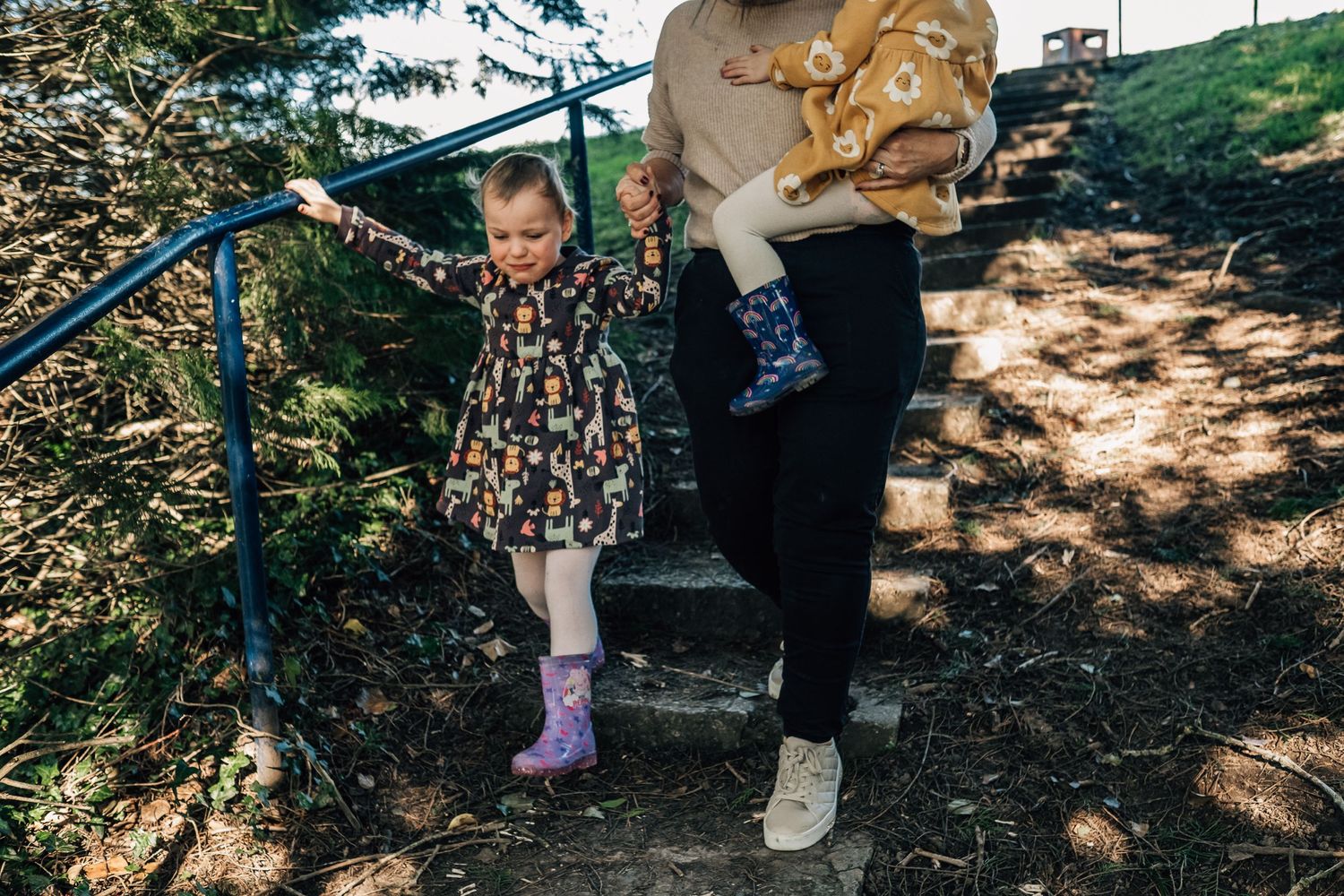 A young child in a floral dress and purple boots walks down outdoor steps with an adult on a nature path.