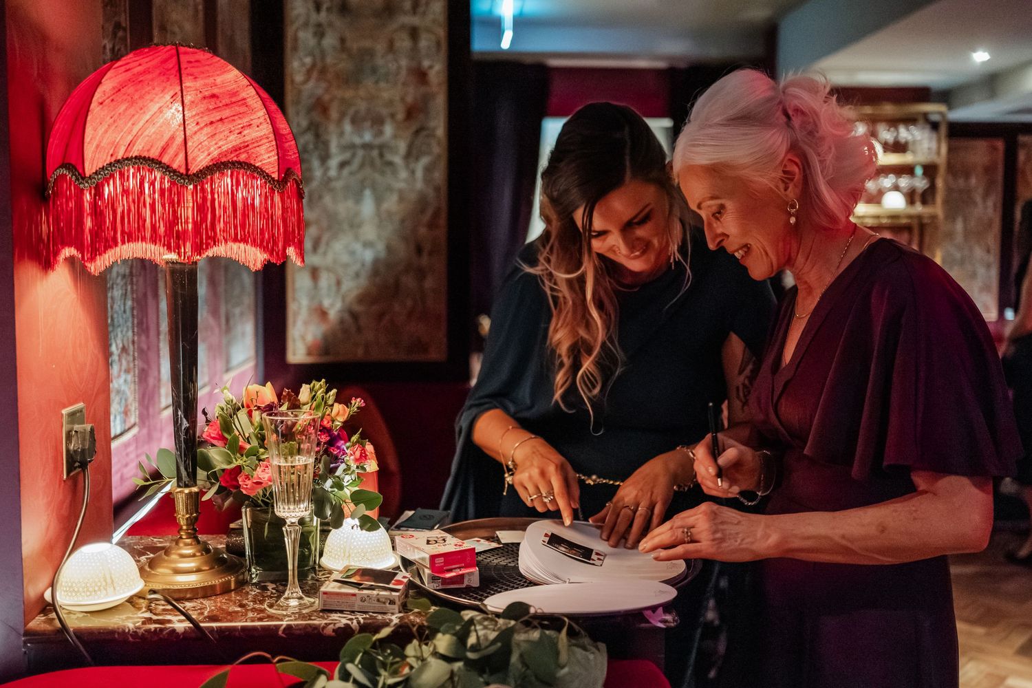 A vintage-style bar scene with a red fringed lampshade and two people examining papers on a table.
