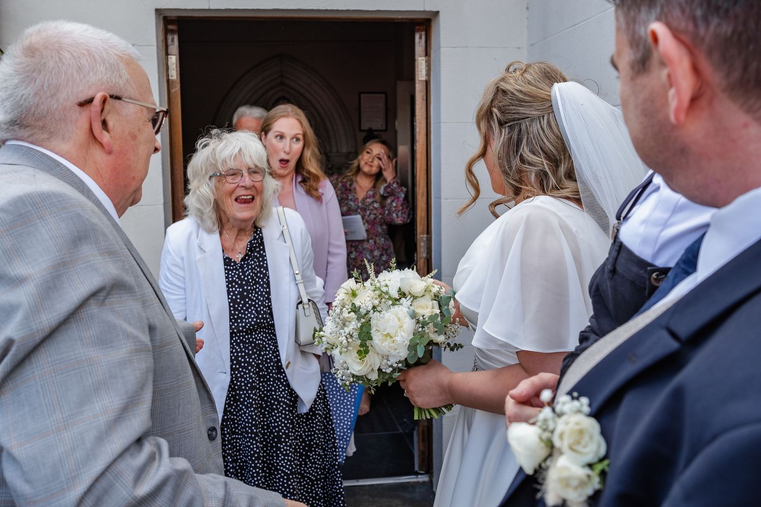 Wedding guests gather at a church doorway holding white flower bouquets.
