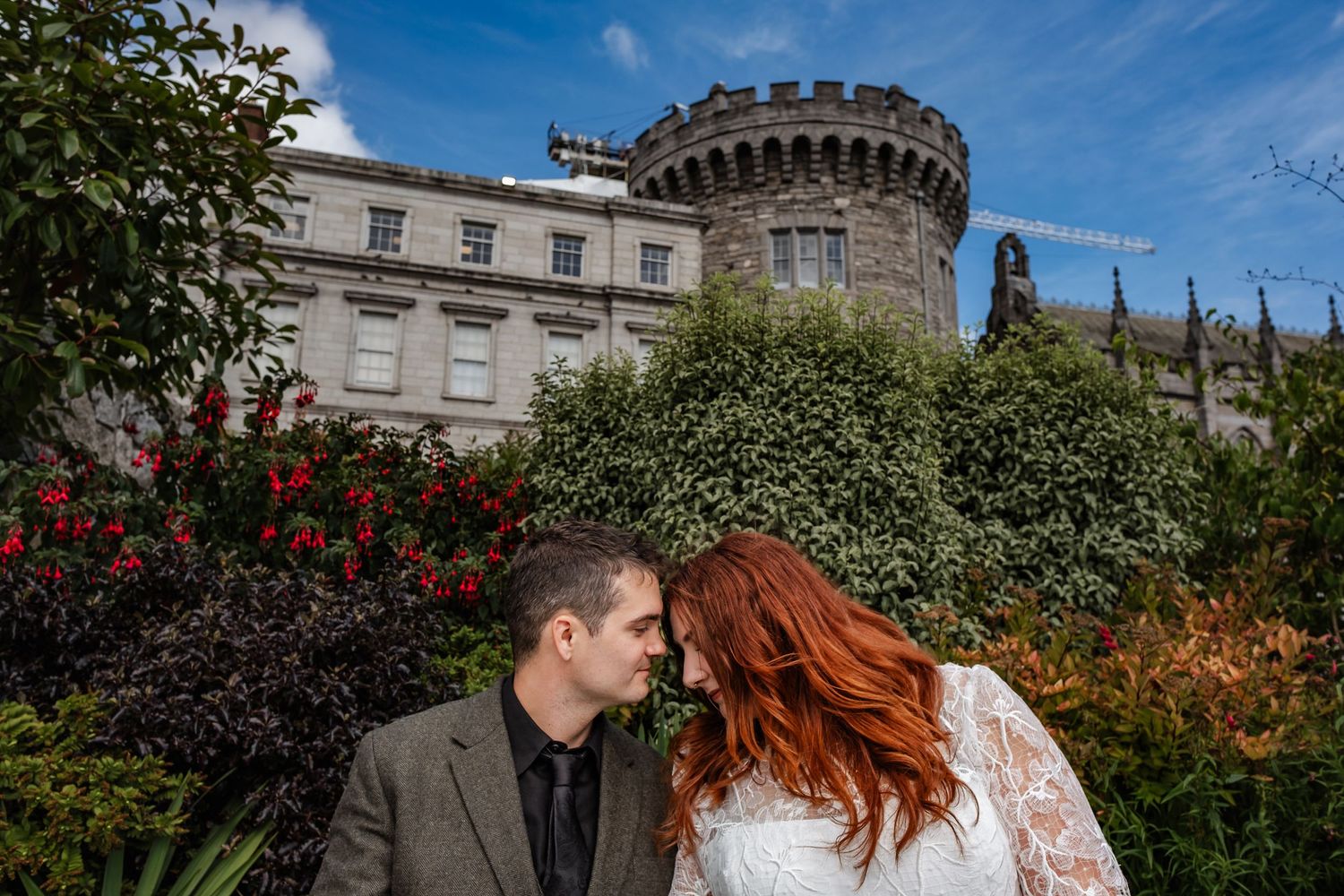 A couple poses intimately near a castle tower surrounded by blooming red flowers and greenery.