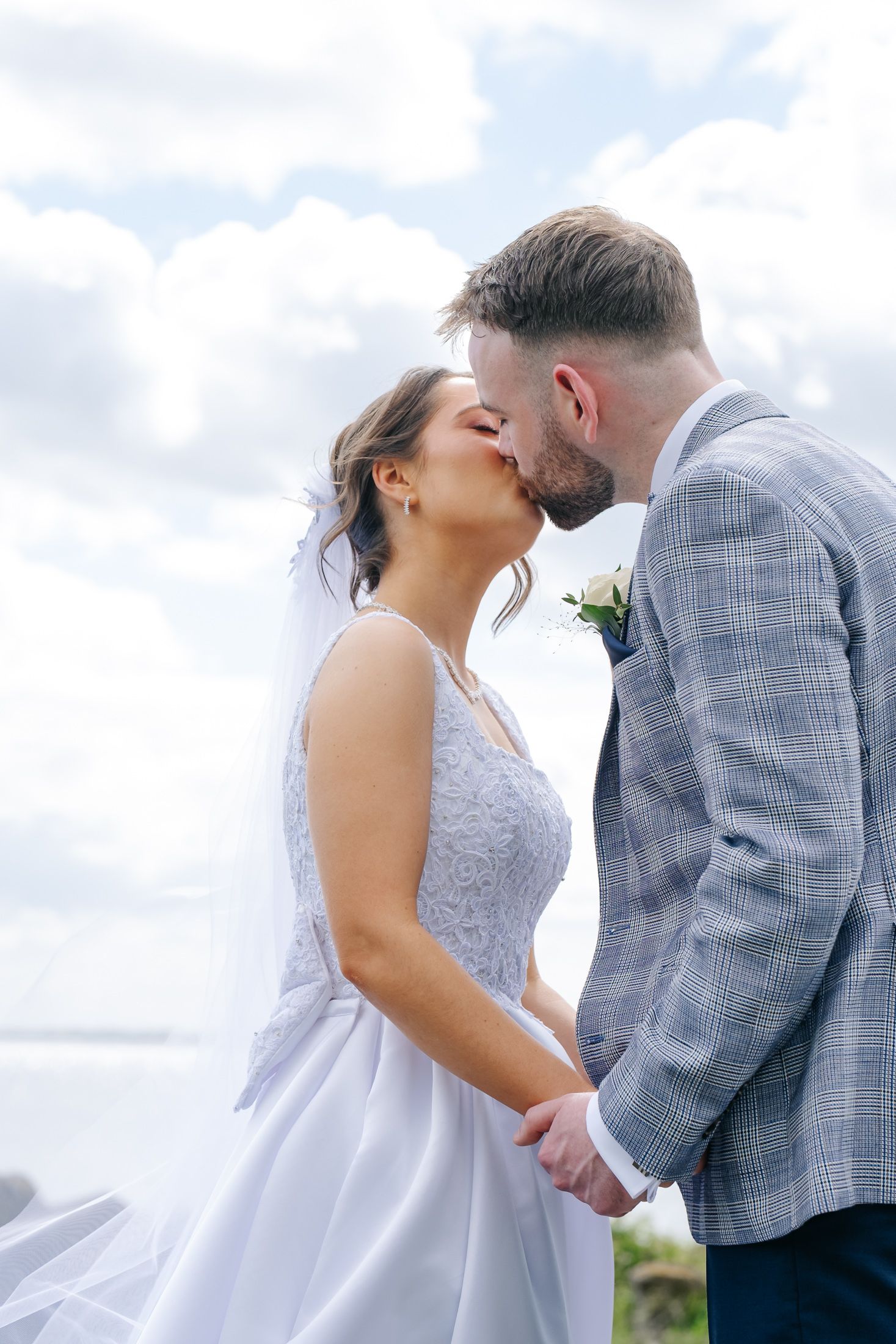 Bride and groom share a romantic kiss outdoors, holding hands, with a cloudy sky backdrop on their wedding day.