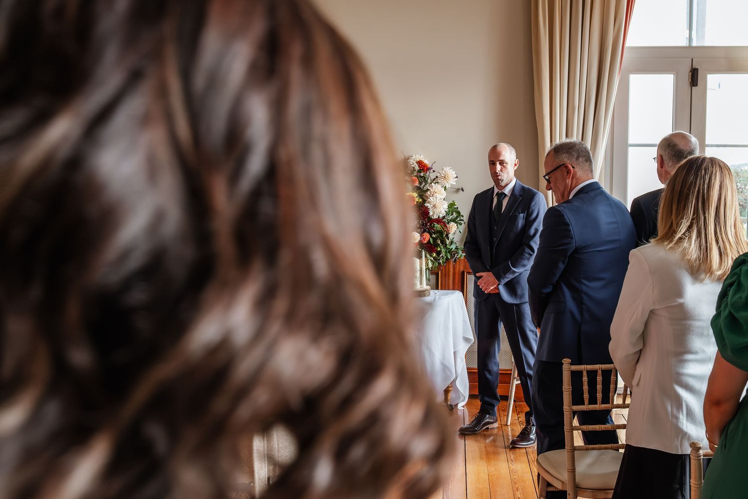 Wedding guests gather around the groom during an indoor ceremony with floral arrangements.
