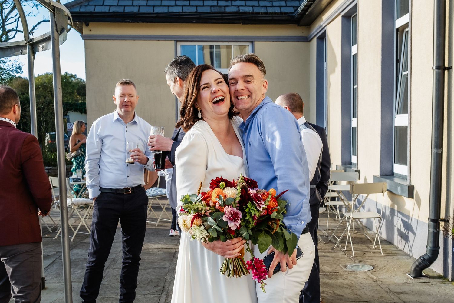 Joyful bride holding a colorful bouquet embraces a laughing guest at an outdoor wedding reception.