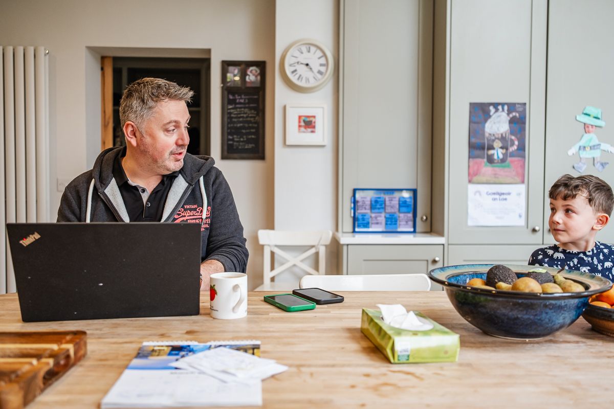 Man working on laptop at kitchen table with young boy sitting beside him, bowl of fruit and papers nearby.