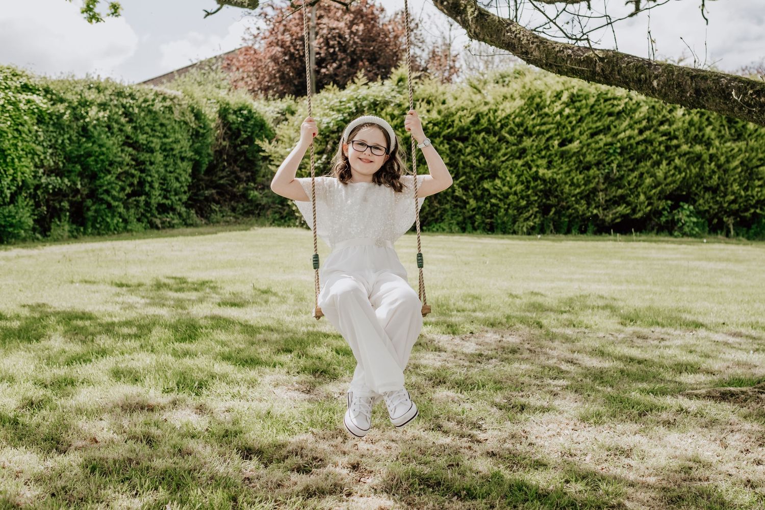 Young girl with glasses sitting on a rope swing in a lush green garden on a sunny day.