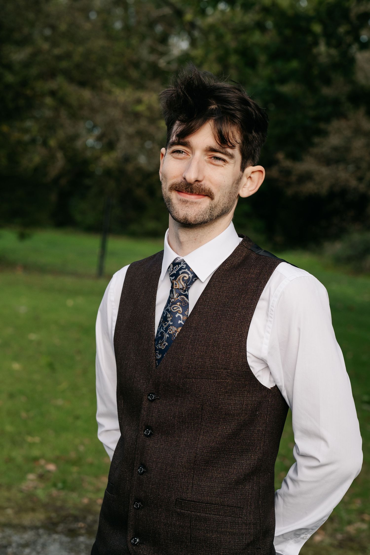 Young man with mustache wearing brown tweed vest, white shirt, and paisley tie, smiling outdoors in a green park.