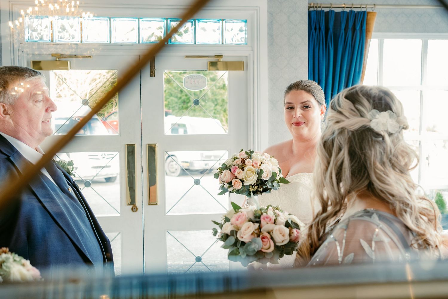 A wedding ceremony moment captured through an architectural frame with blue curtains and floral bouquets.