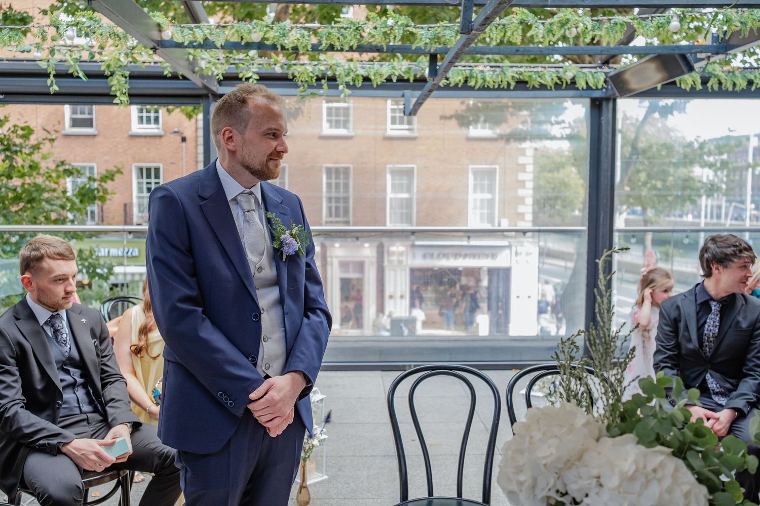 People in formal attire gathered at an outdoor venue with tables and chairs on a patio.
