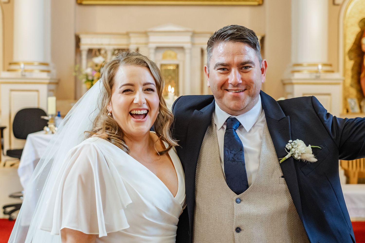 A happy newlywed couple smiling together in their wedding attire inside an ornate venue.