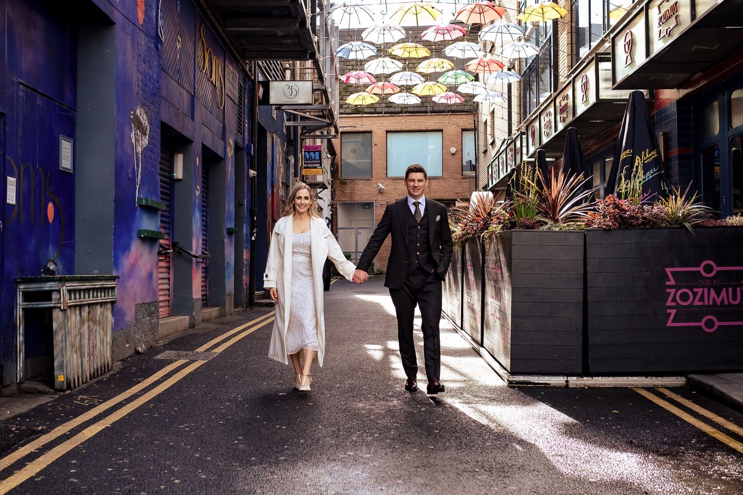 Couple walking hand in hand under colorful umbrellas in an urban alley.