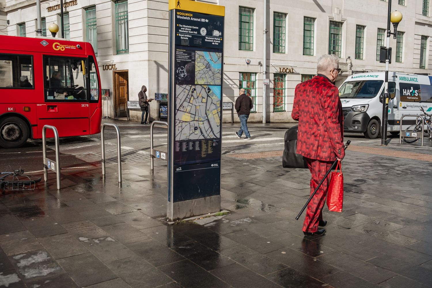 Person in bright red outfit walking on rainy street with red bus in background.