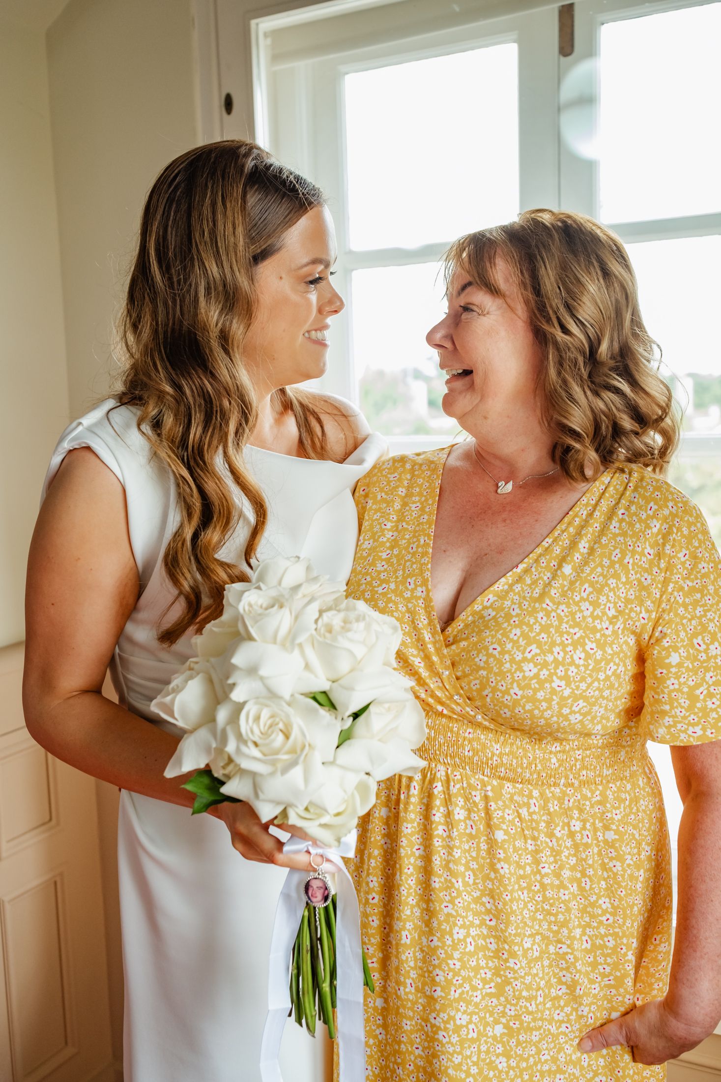 Two women in white and yellow dresses share a joyful moment with white roses.